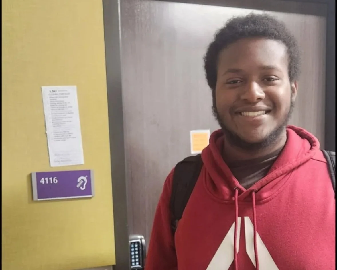 A young man with a backpack smiling in front of an elevator with the room number 4116.