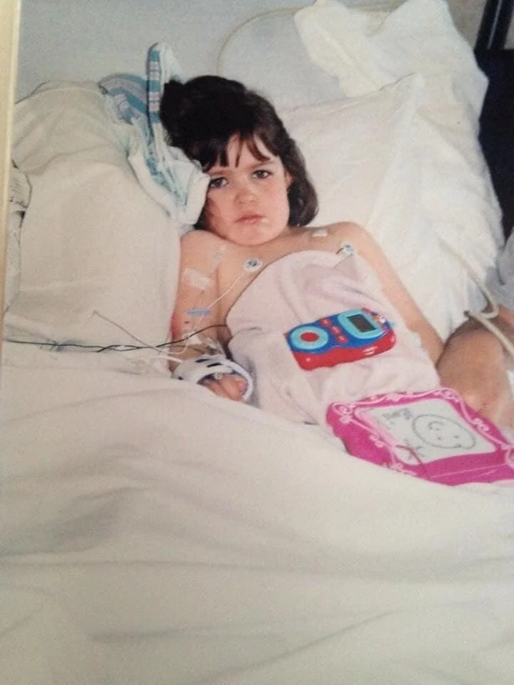 Young girl lying in a hospital bed with medical wires, a pink toy tablet, and a small pink drawing pad.