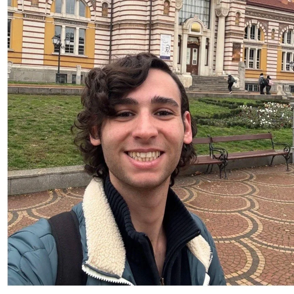 A young man with wavy brown hair smiling for a selfie in front of a historic brick building with stone columns and steps, a grassy area, flowerbeds, and park benches.