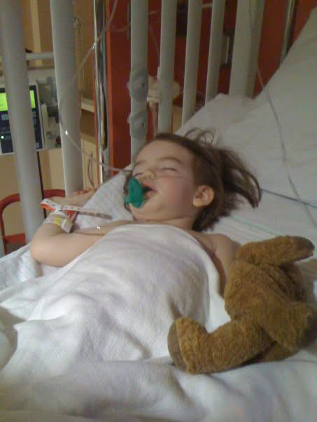 A young girl lying in a hospital bed with a nasal cannula, holding a stuffed animal, and smiling.
