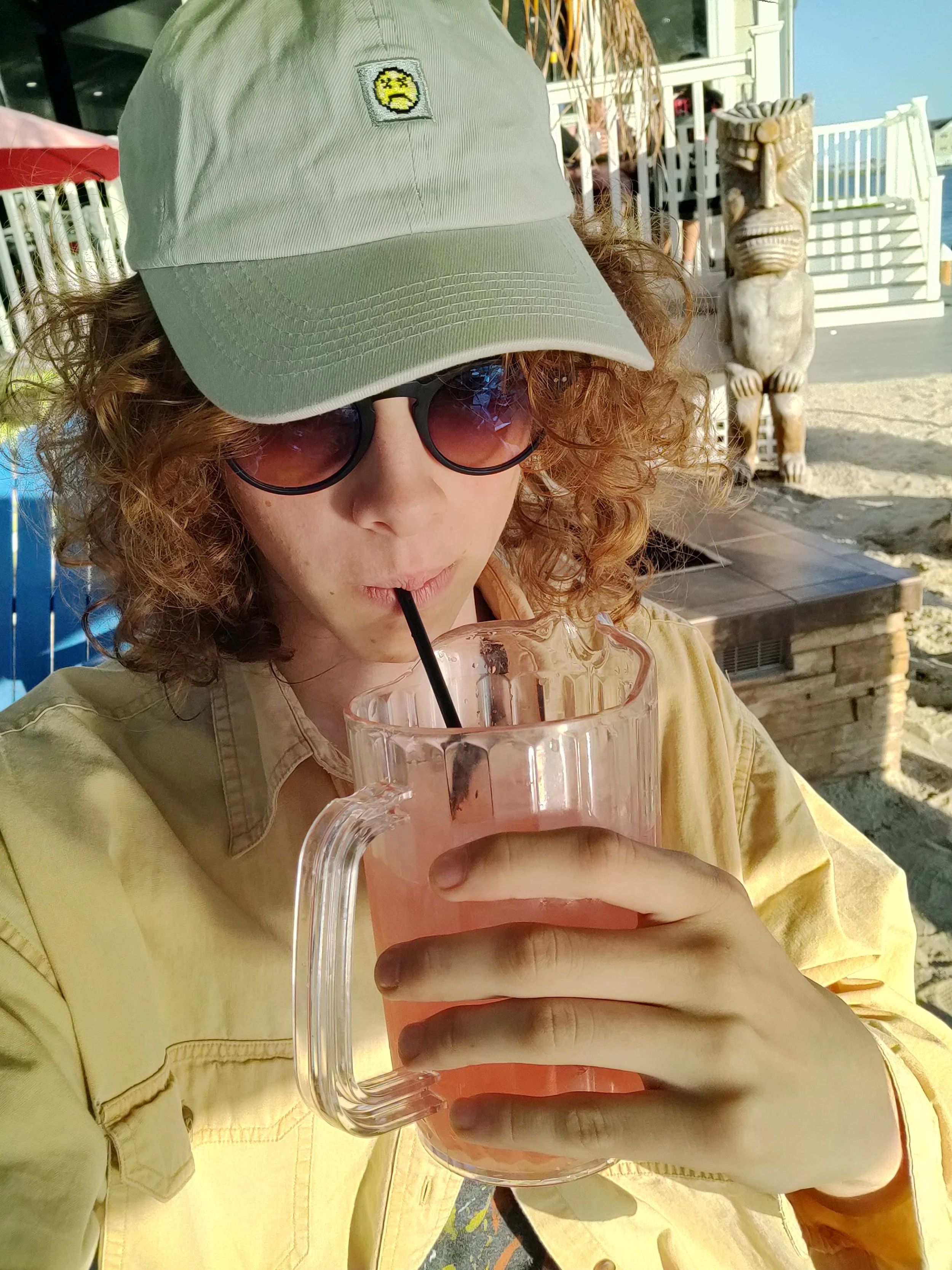 Person with curly hair wearing a beige cap with a sad emoji patch, sunglasses, and a yellow shirt, drinking a pink beverage through a straw at an outdoor location with tribal art and a railing in the background.