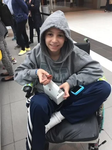 A young boy in a wheelchair wearing a gray hoodie and navy pants, smiling and holding a white box with a blue electronic device nearby, in a public place with other people in the background.