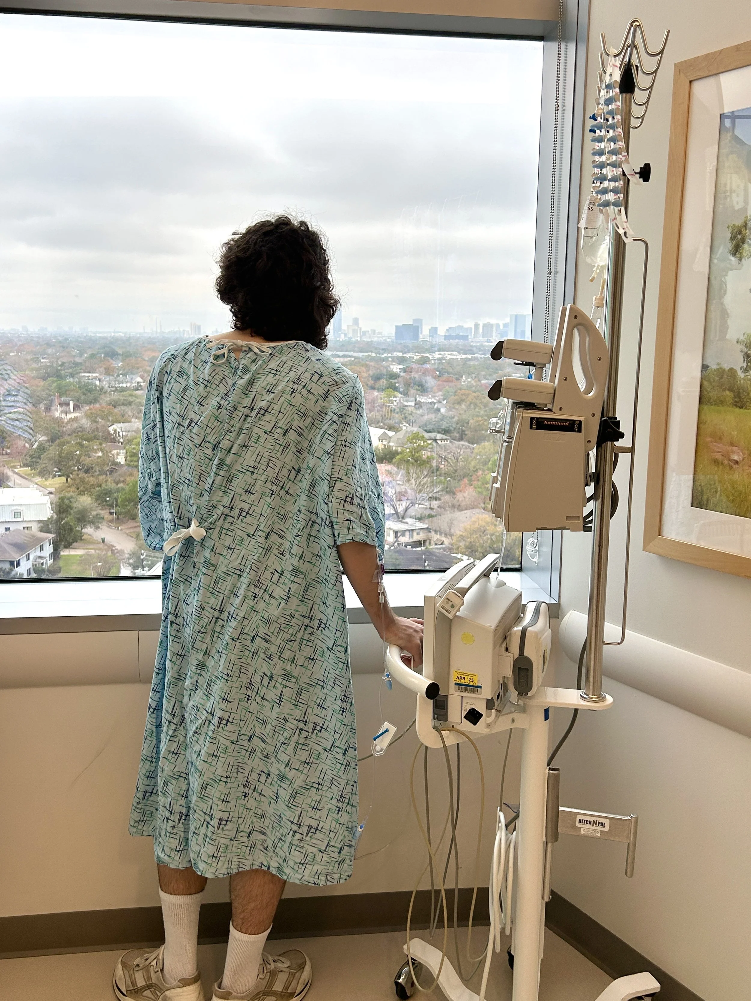 A person in a hospital gown standing by a window, with medical equipment such as IV lines and monitors nearby, overlooking a cityscape with trees, buildings, and cloudy skies.