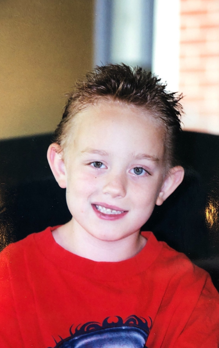 A young boy with short spiky blond hair, blue eyes, and a big smile wearing a red shirt, sitting indoors on a black chair.