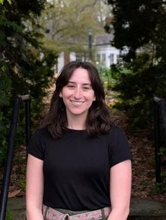 Young woman with shoulder-length dark hair smiling outdoors in a park or garden setting.