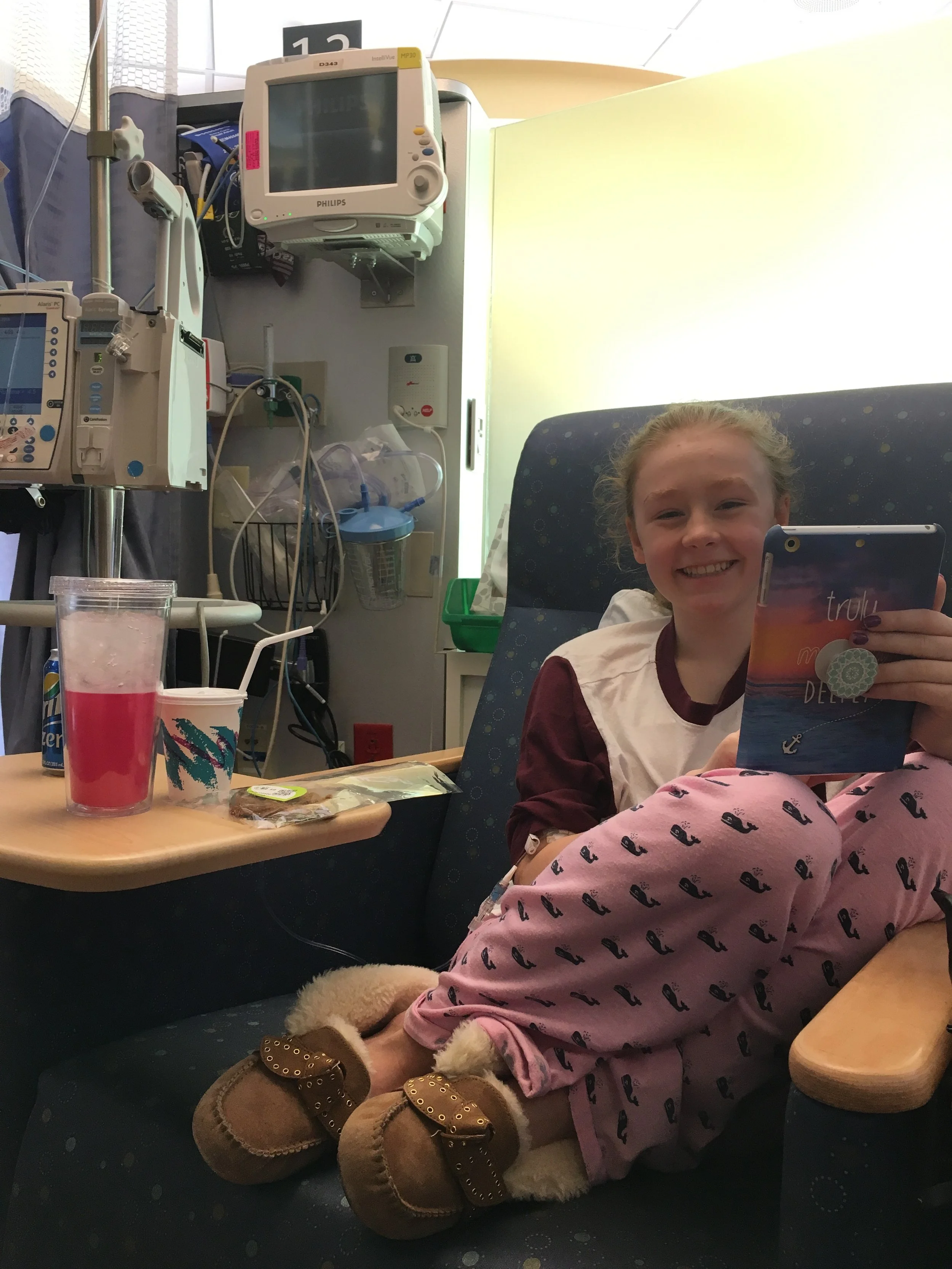 A young girl smiling in a hospital bed, taking a selfie with her smartphone, wearing pink pajamas with black whale prints and brown slippers. Medical equipment and monitors are visible in the background.
