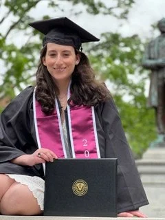 A young woman in graduation cap and gown holding a diploma and sitting outdoors in front of a statue.