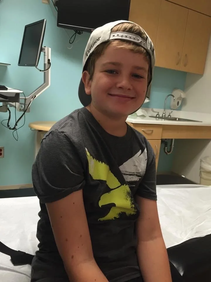 A young boy with a smile, wearing a baseball cap backward and a Batman T-shirt, sitting on a hospital bed in a medical room.