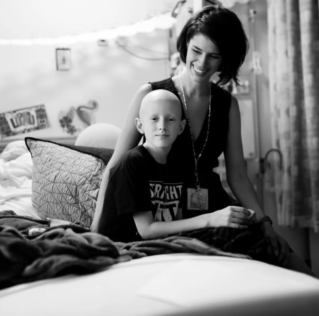 A woman and a young boy sitting on a bed, smiling and looking at the camera in a room with a decorated wall.