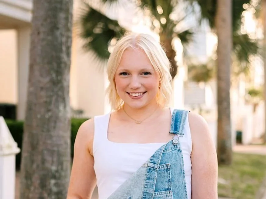 A young woman with blonde hair smiling outdoors, wearing a white tank top and denim overalls, with palm trees in the background.