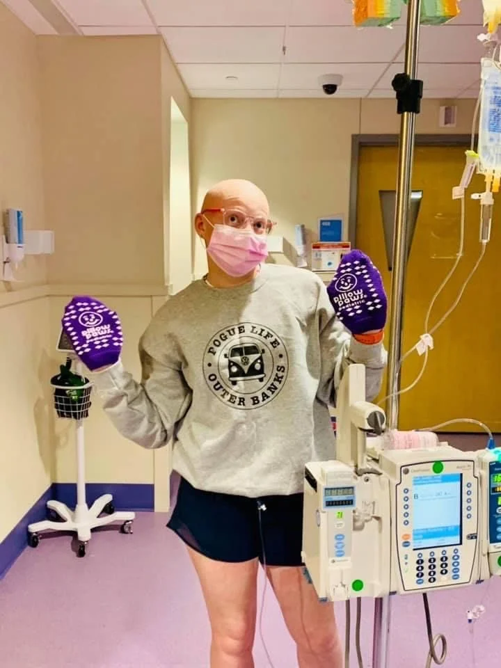A woman in a hospital room wears a pink face mask, glasses, a grey sweatshirt, and purple gloves. She stands in front of IV equipment and a bear-shaped IV pole, striking a playful pose with her arms out.