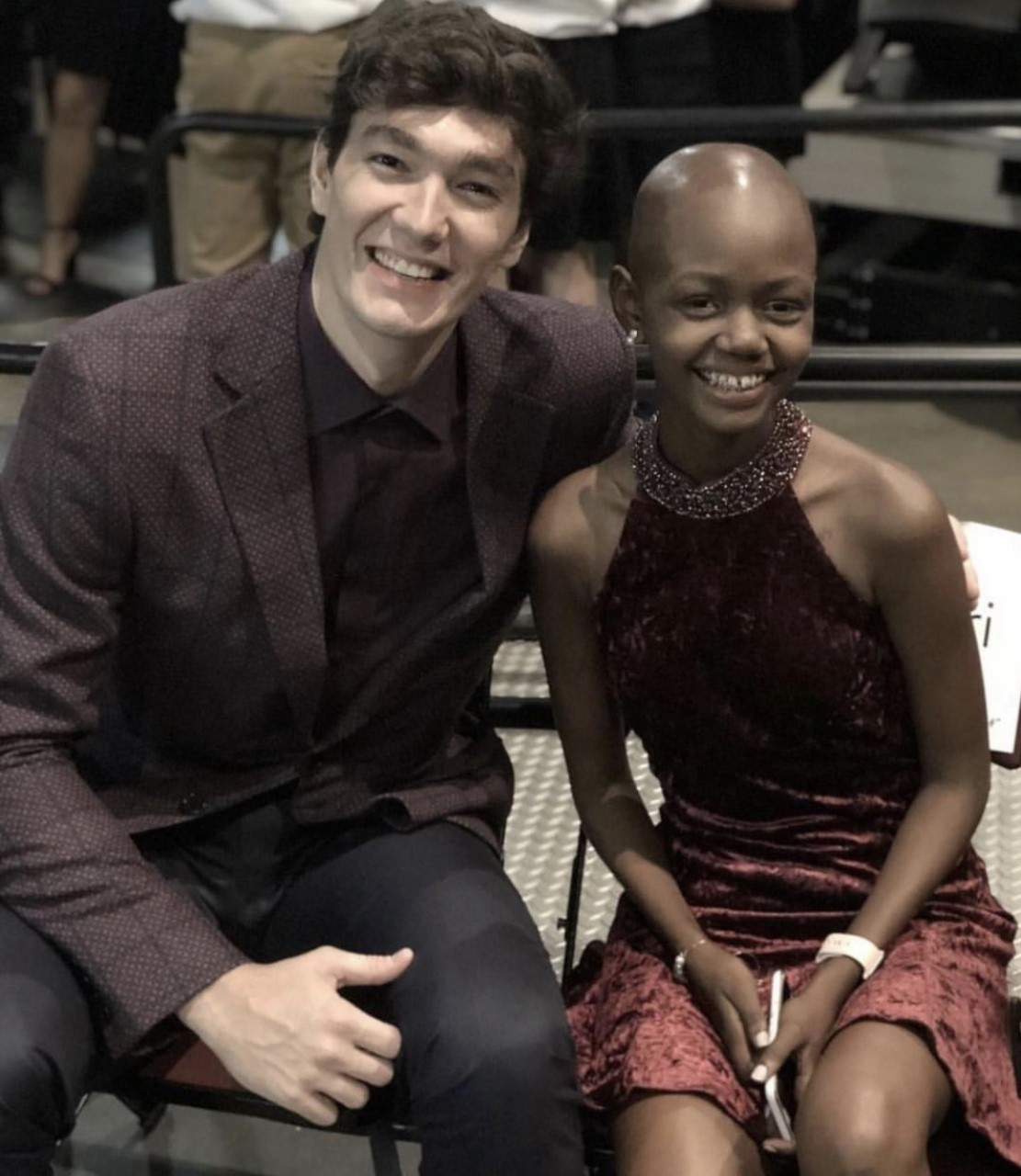 A man and a woman sitting indoors, smiling at the camera, at a formal or semi-formal event.