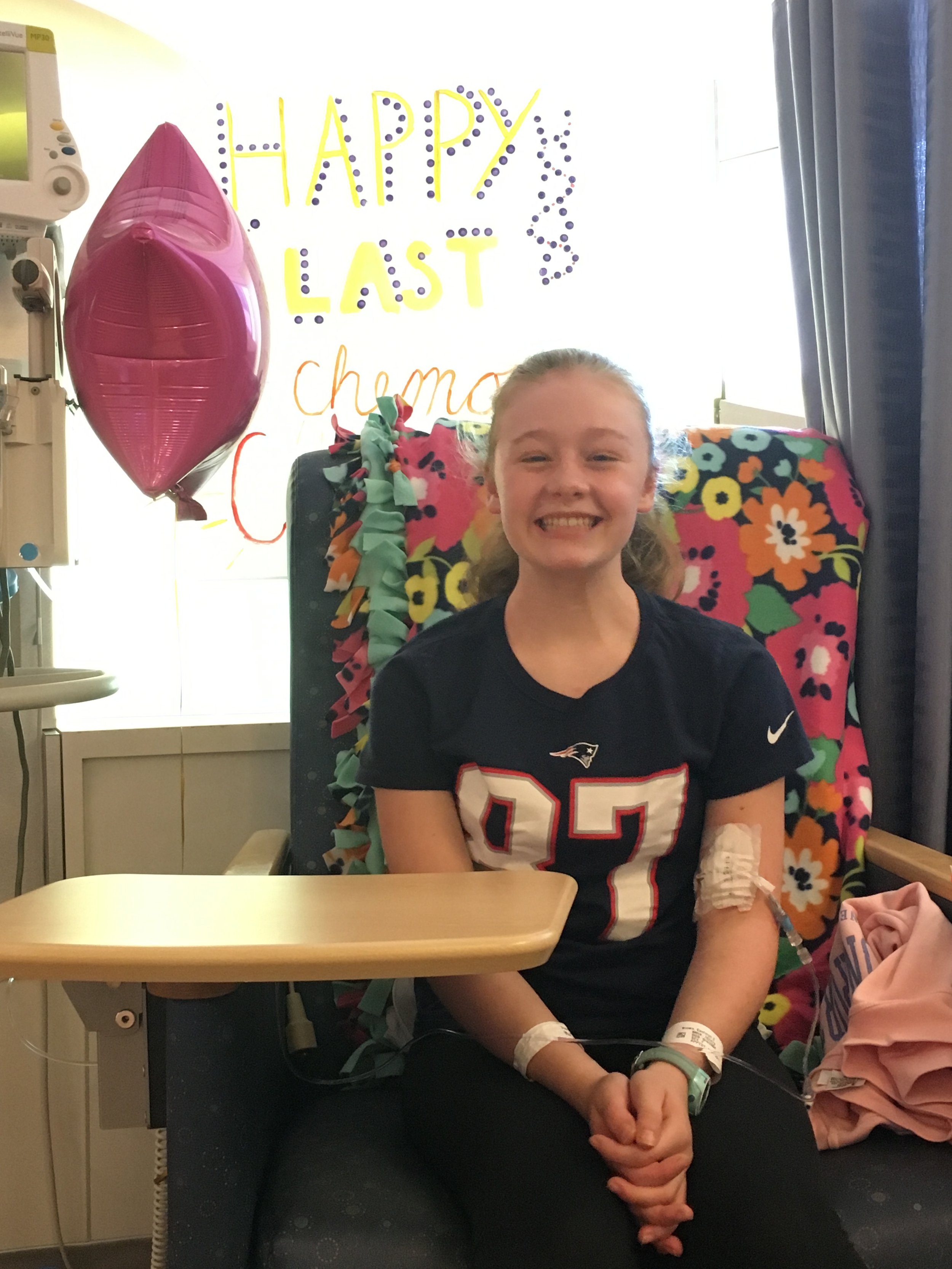 A young girl sitting in a hospital bed, smiling with an IV in her arm, wearing a black sports jersey, with festive decoration in the background and a balloon.