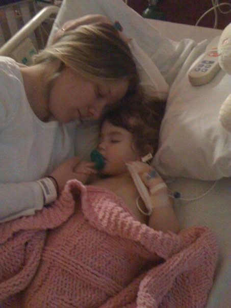 A woman sleeping next to a young girl with a nasal oxygen tube, lying in a hospital bed covered with a pink blanket.