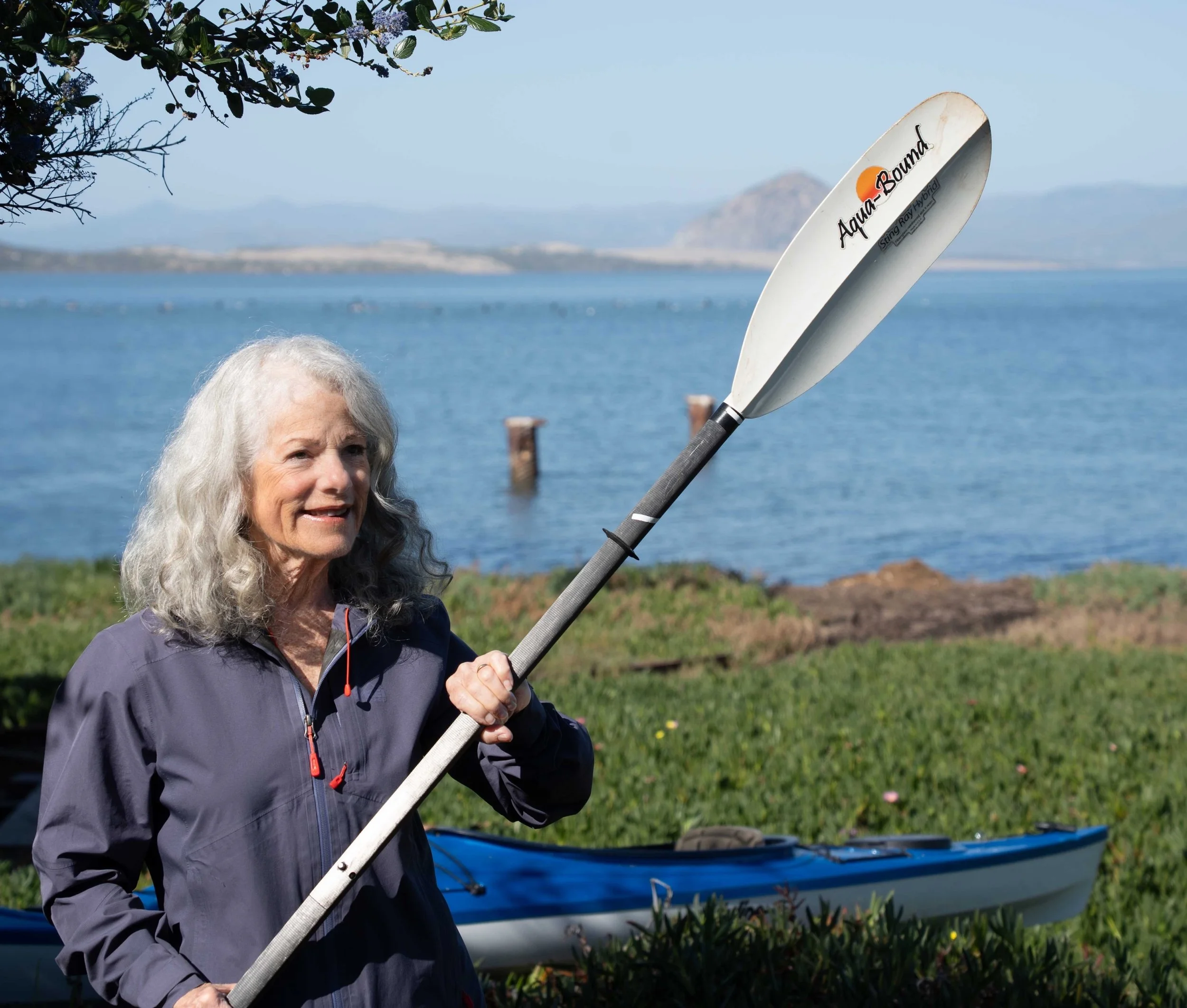 The author Sharon Sheltzer holding an oar with a kayak and a bay in the background.