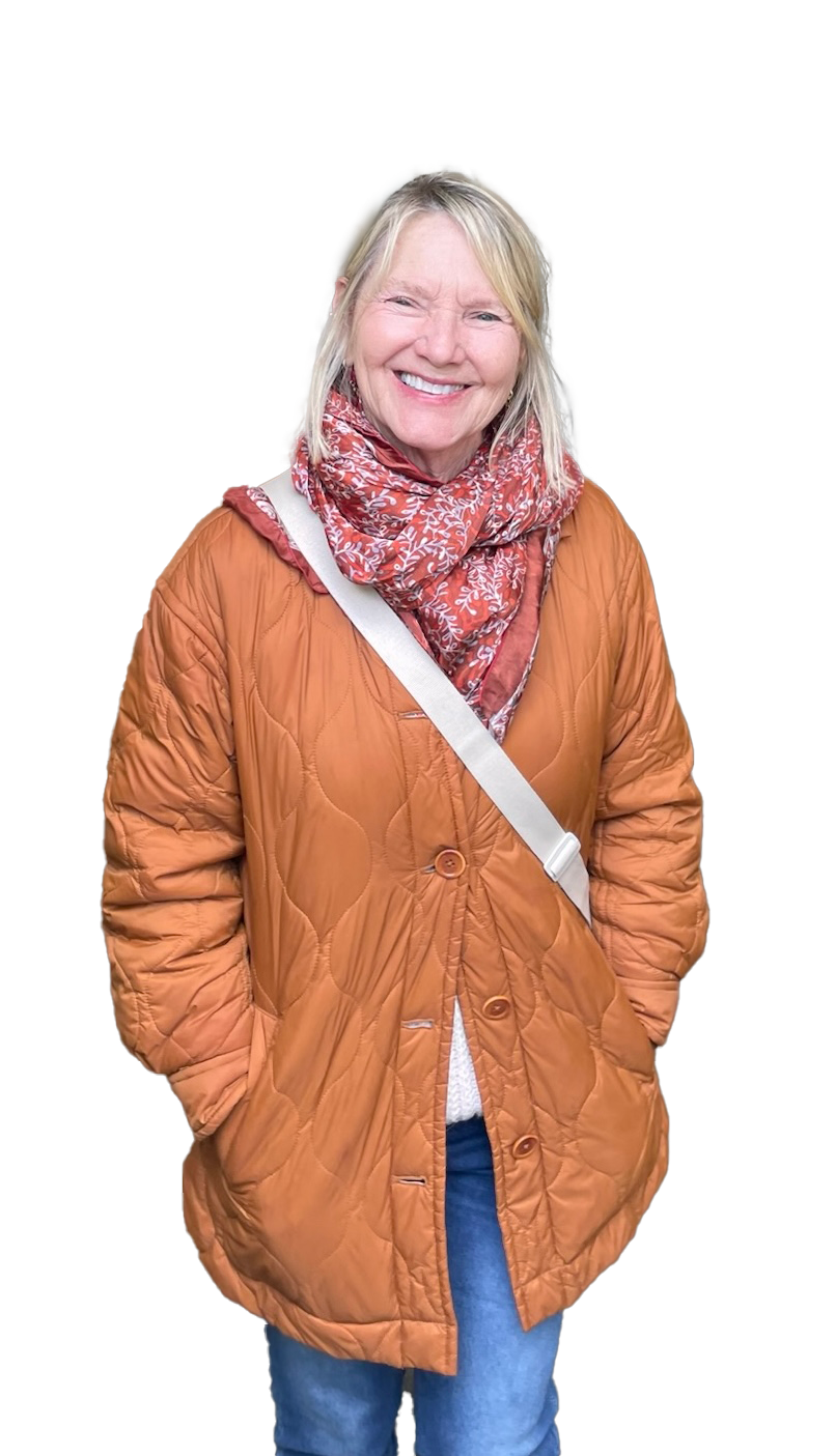 A woman stands smiling wearing a pumpkin colored quilted jacket and a cross the body purse.