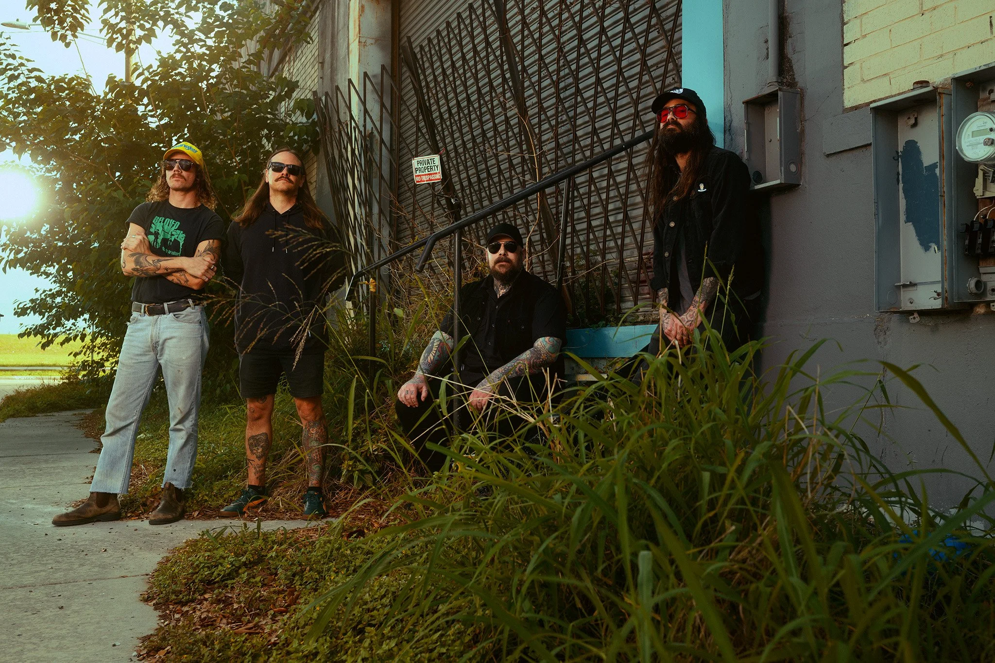Four men with long hair and tattoos posing outdoors near a building with a metal gate and a sign that reads 'Private Property No Trespassing'. One man is wearing sunglasses, another is wearing a yellow cap, and they are dressed in casual dark clothin