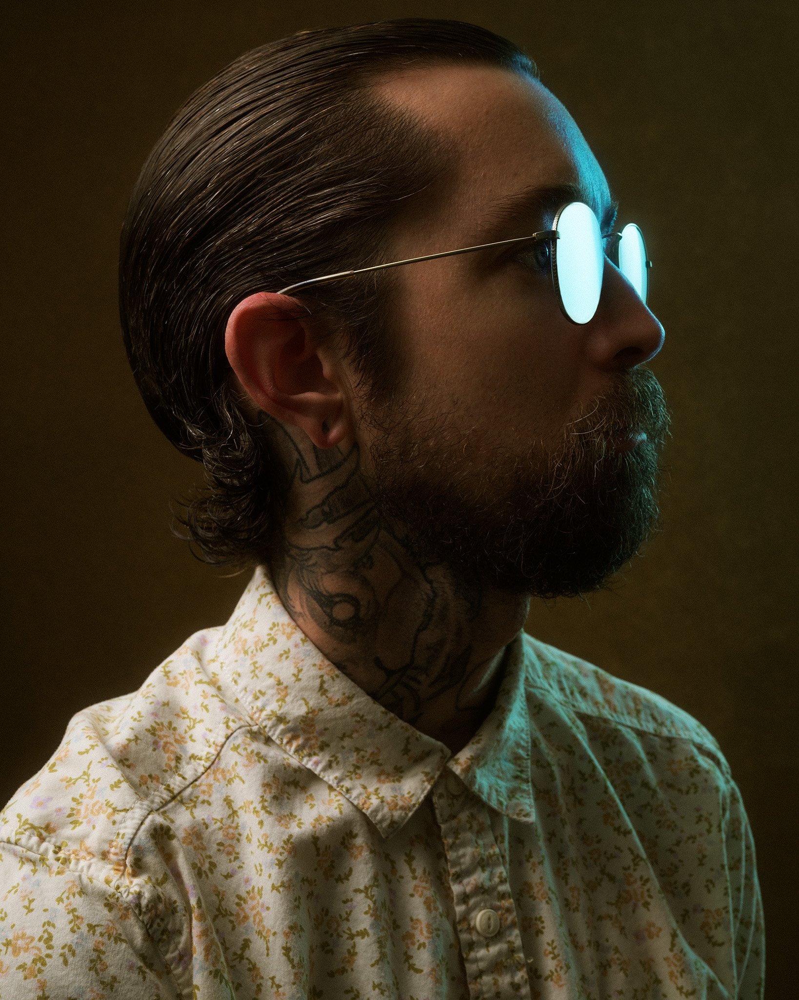 A side profile of a man with slicked-back dark hair, wearing round glasses with bright reflections, a mustard-colored beard, and a floral shirt with yellow and pink patterns, against a dark background.