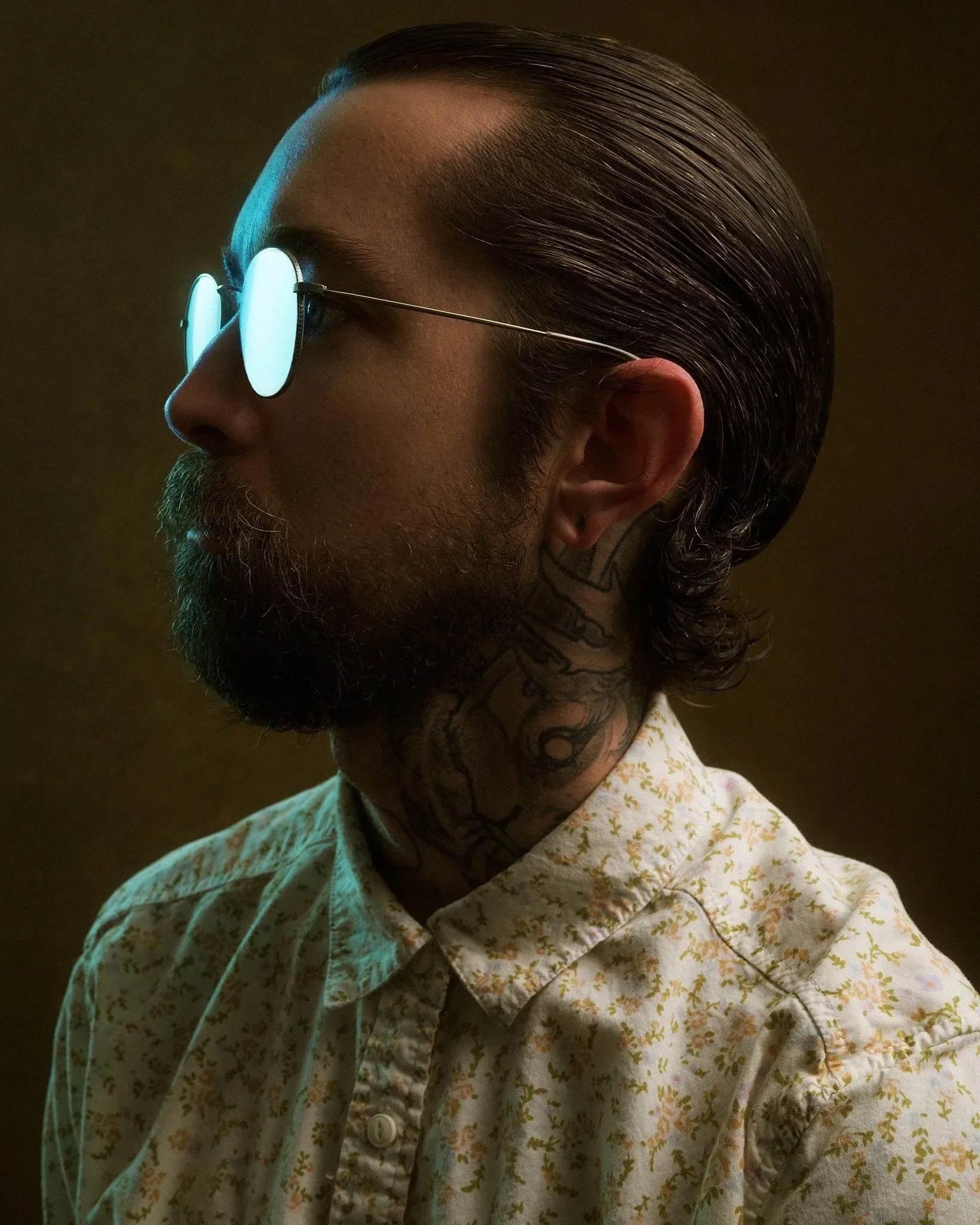 Side view of a man with dark, slicked-back hair, wearing round reflective sunglasses and a light-colored shirt with a floral pattern.