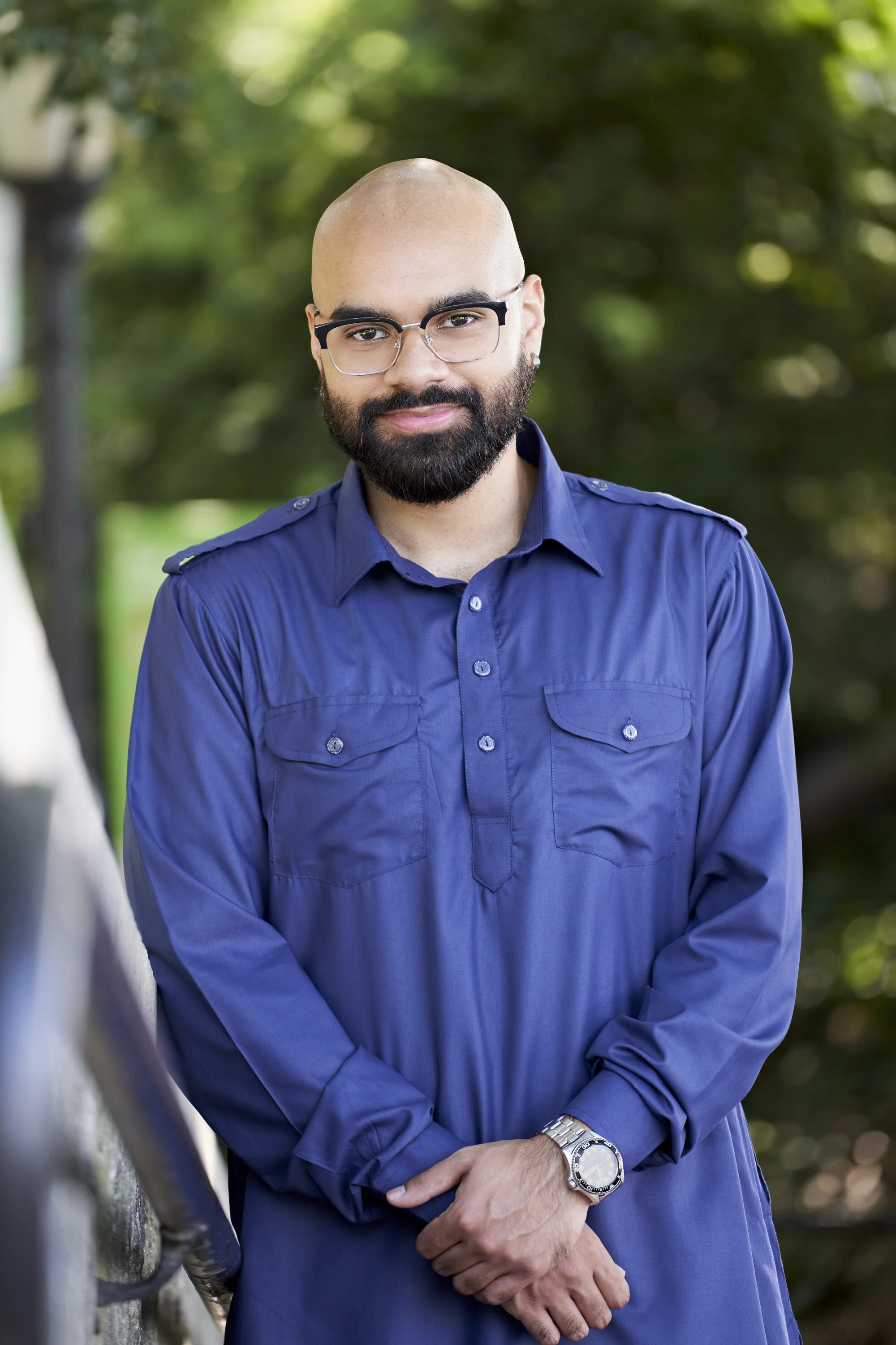 A bald man with glasses, a beard, and wearing a blue shirt standing outdoors with trees in the background.