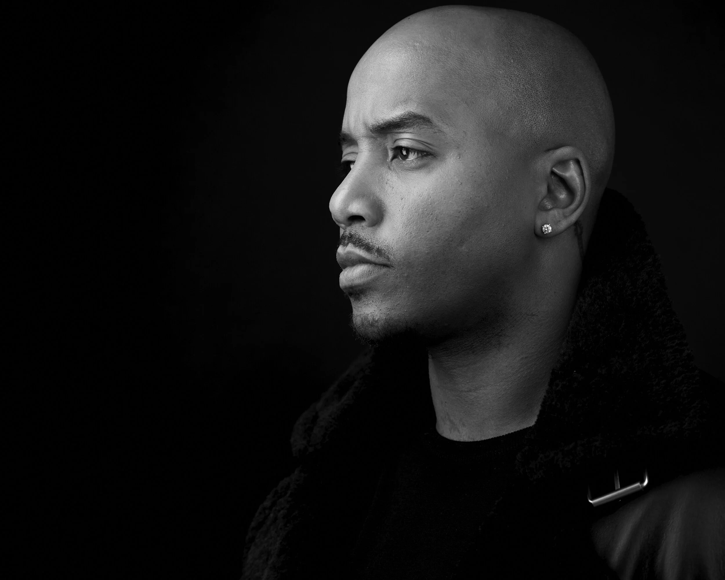 Black and white portrait of a man with a shaved head, earring, and a serious expression, wearing a jacket with a large collar, looking to the left.