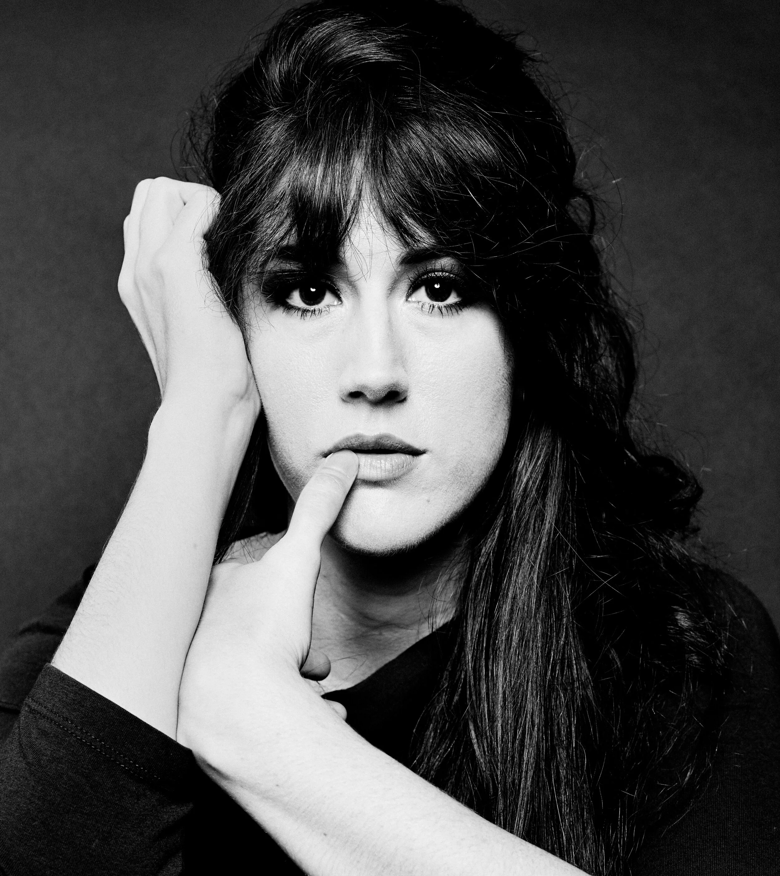 Black and white portrait of a woman with dark hair and expressive eyes, with one hand resting on her face and the other touching her lips.
