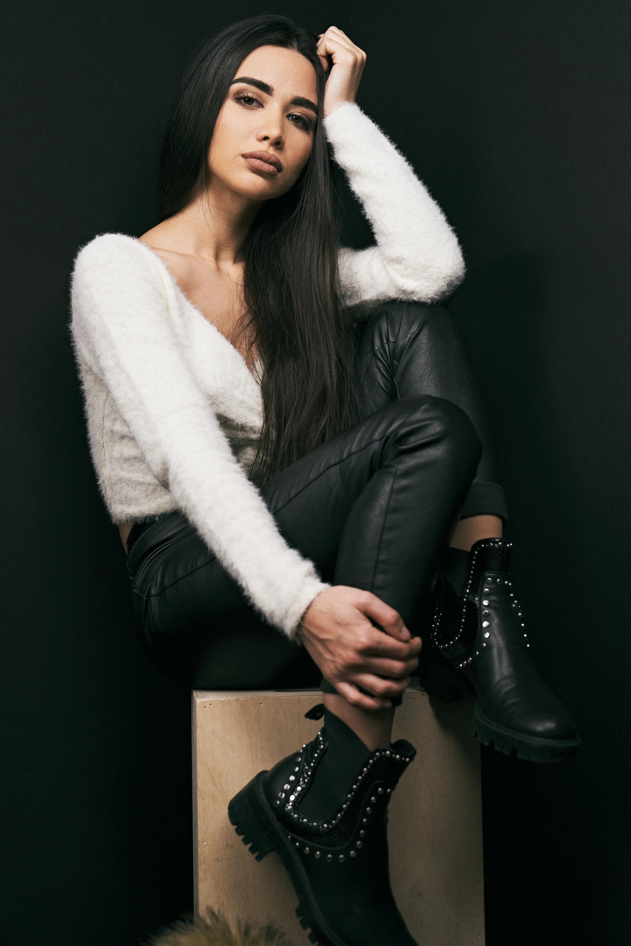 A woman with long dark hair, wearing a white fuzzy sweater, black leather pants, and black studded combat boots, poses sitting on a wooden box against a dark background.