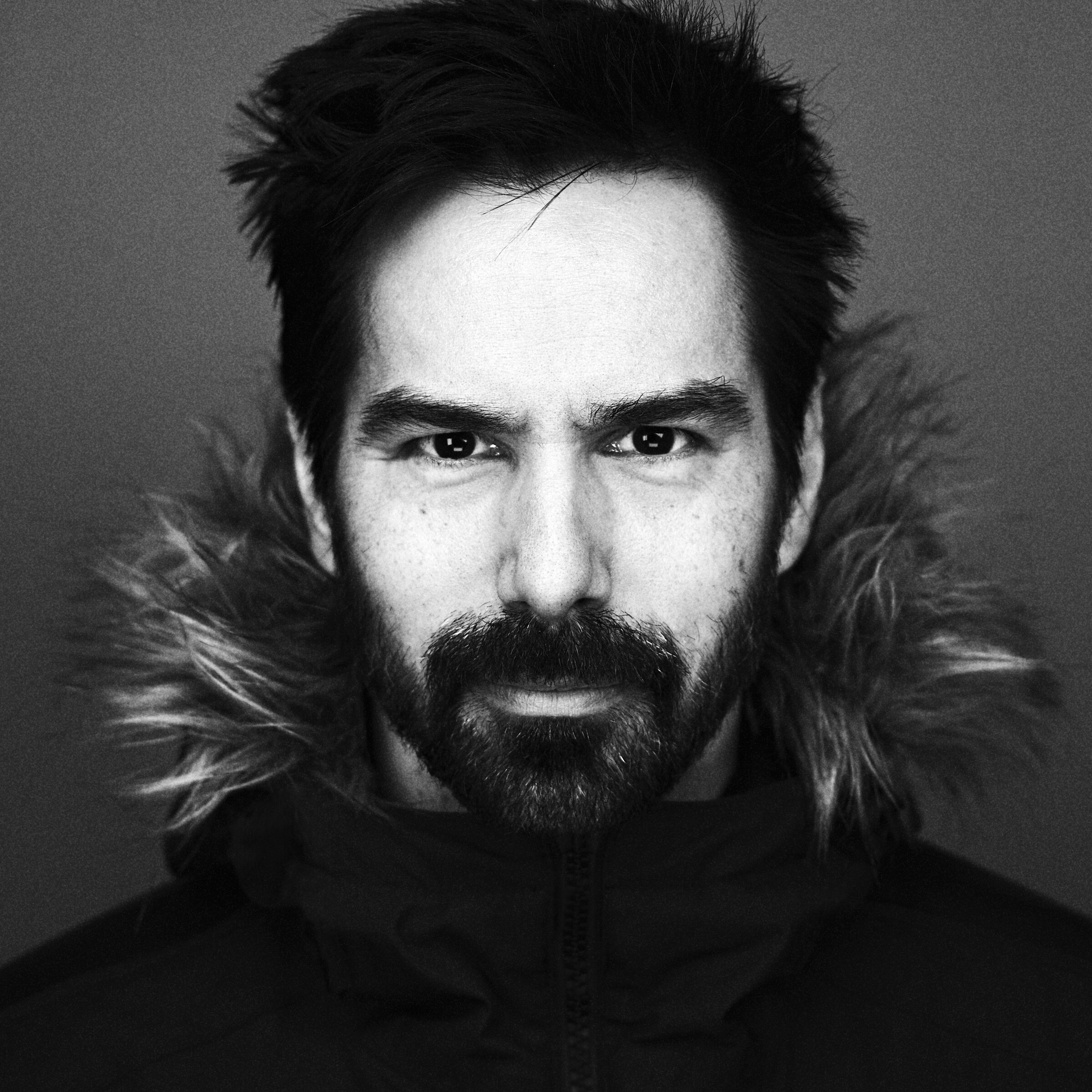 Black and white portrait of a man with a beard and mustache wearing a jacket with a fur-lined hood, looking directly at the camera.