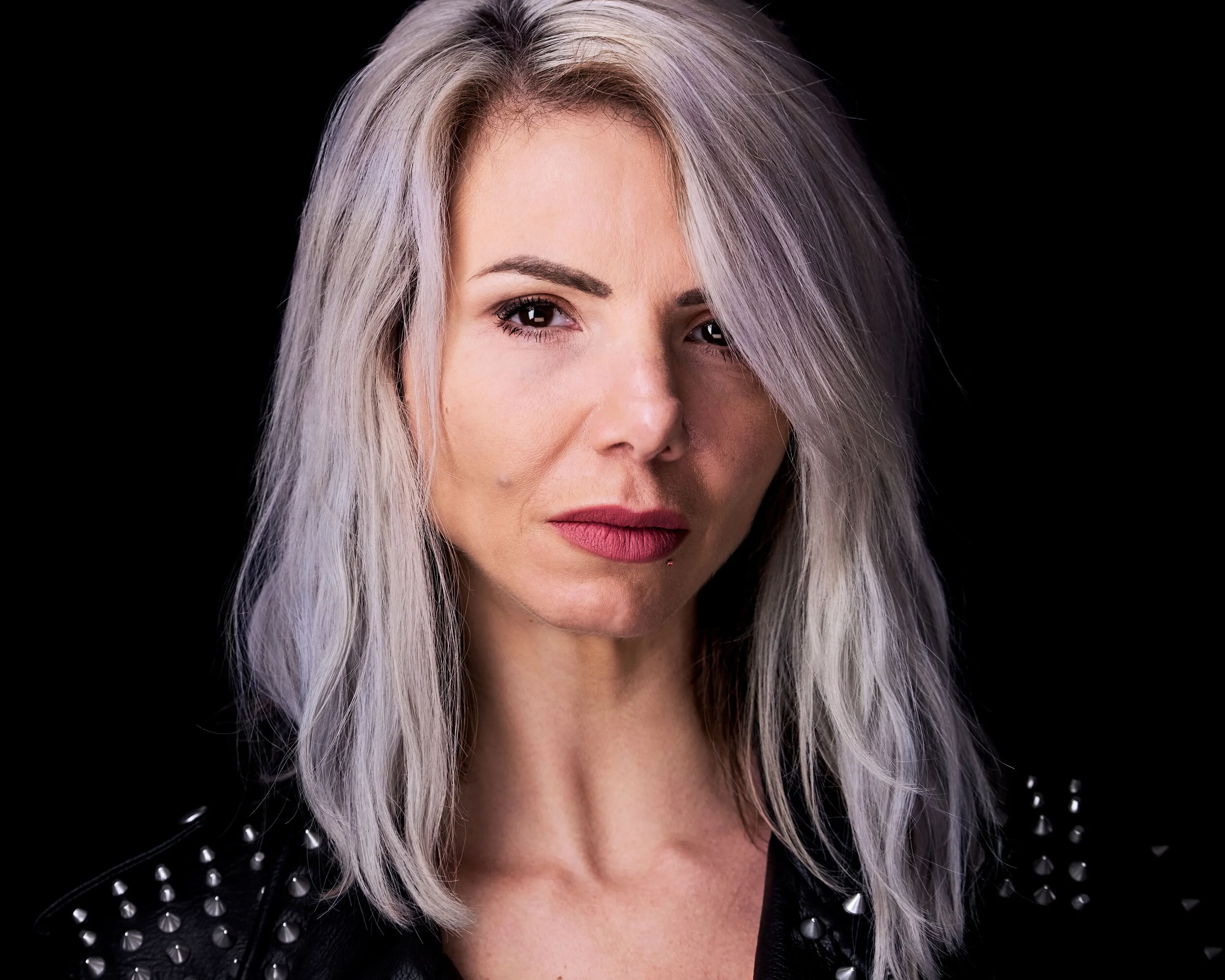 Close-up portrait of a woman with platinum blonde, wavy hair, wearing a black leather jacket with silver studs, against a black background.