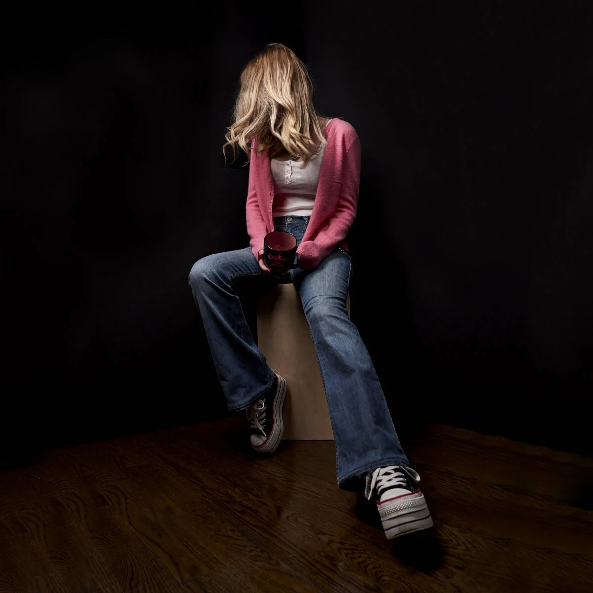 A woman sitting on a wooden block against a black background, wearing a pink cardigan, white top, and blue jeans, holding a black coffee mug.