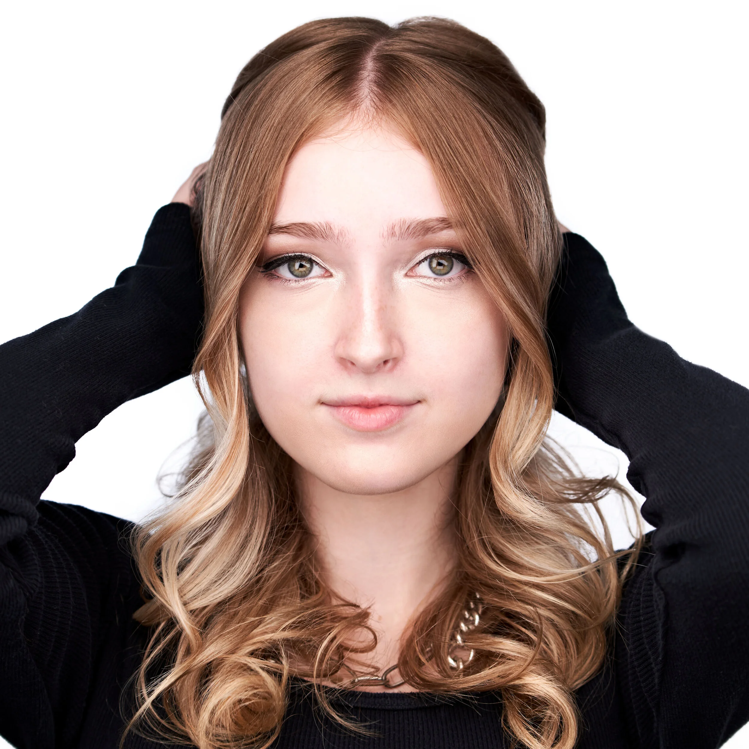A young woman with wavy blonde hair, wearing a black top and a chain necklace, holds her hair back with both hands, looking directly at the camera with a neutral expression.