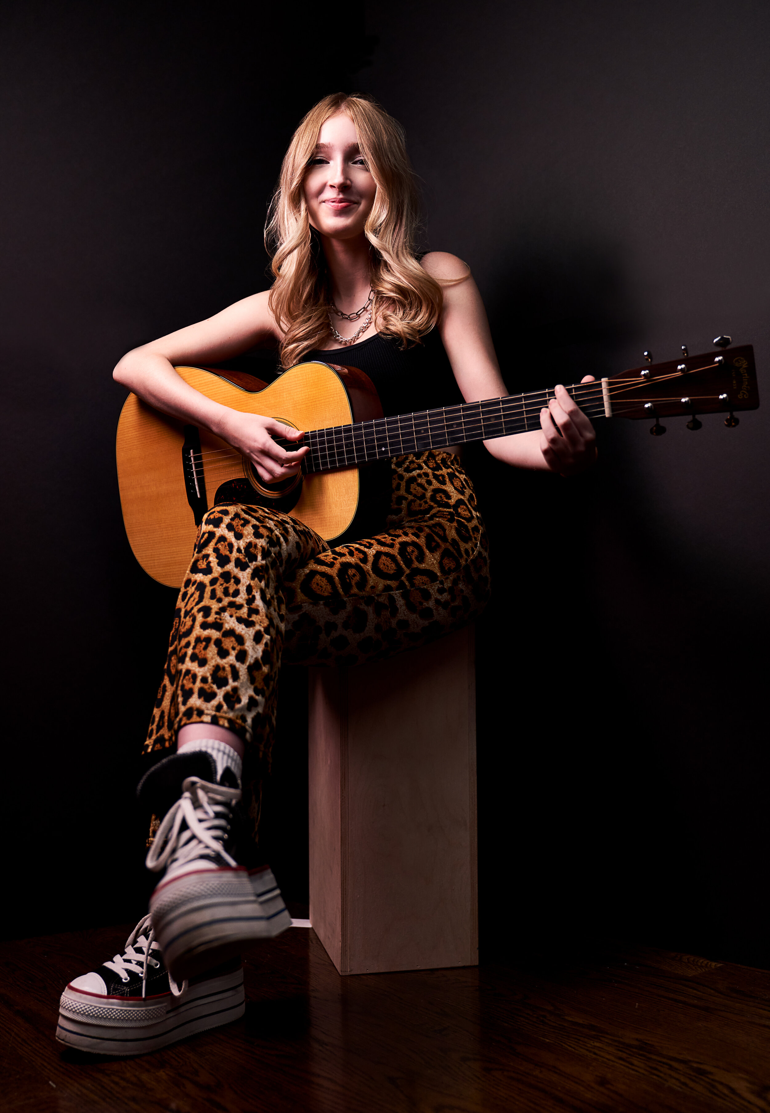 A young woman with light brown hair sitting on a wooden block, playing an acoustic guitar, wearing animal print pants, black top, sneakers, and jewelry, against a dark background.