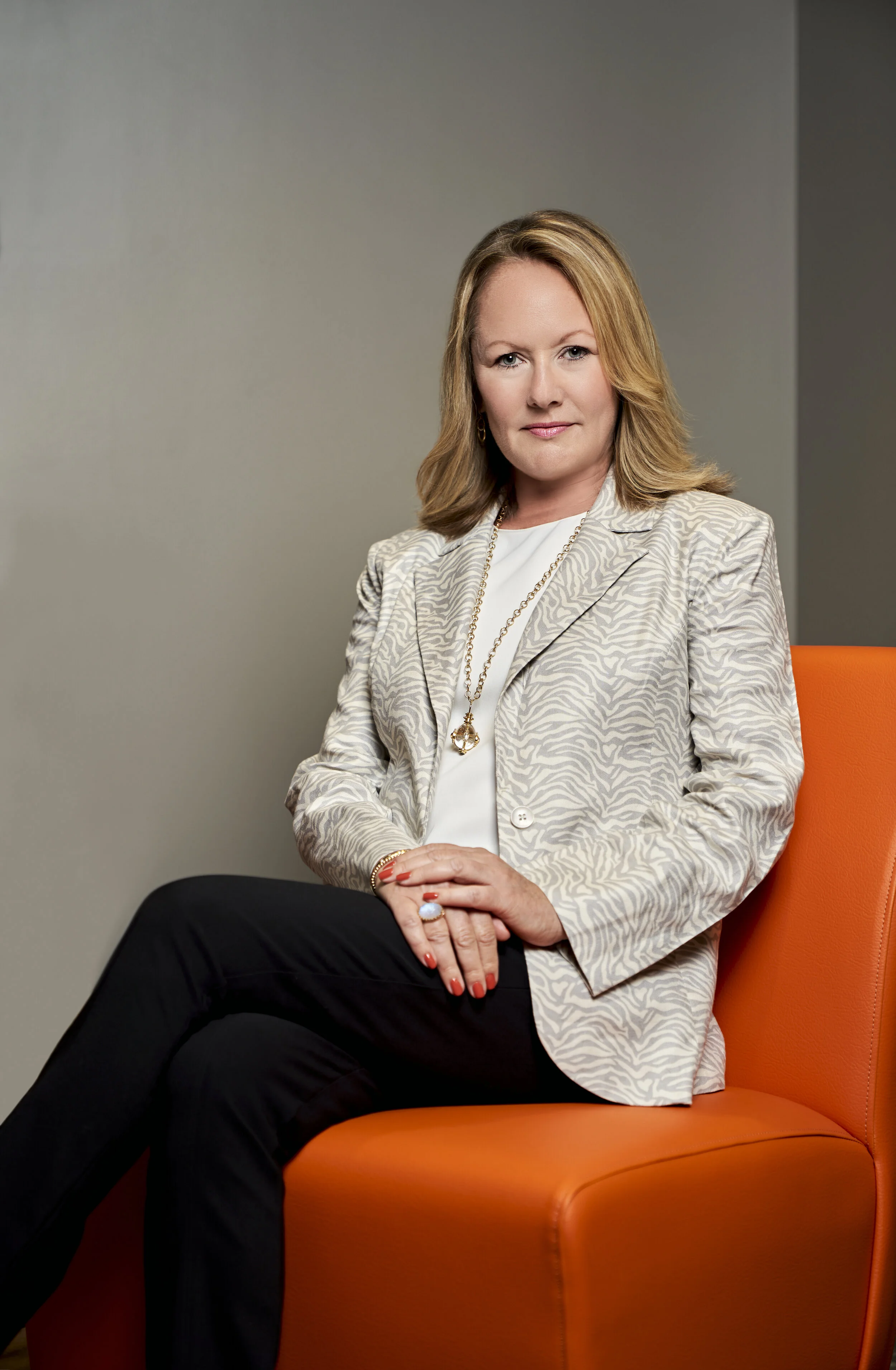 A woman with blonde hair, dressed in a patterned blazer and white top, sitting on an orange chair against a plain gray background, looking confidently at the camera.