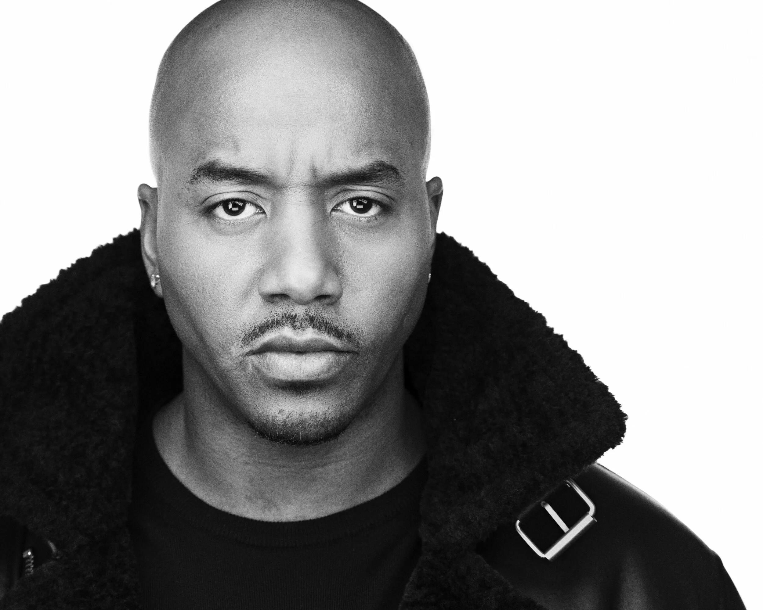 Black and white portrait of a man with a bald head, wearing a coat with a large collar, looking directly at the camera with a serious expression.