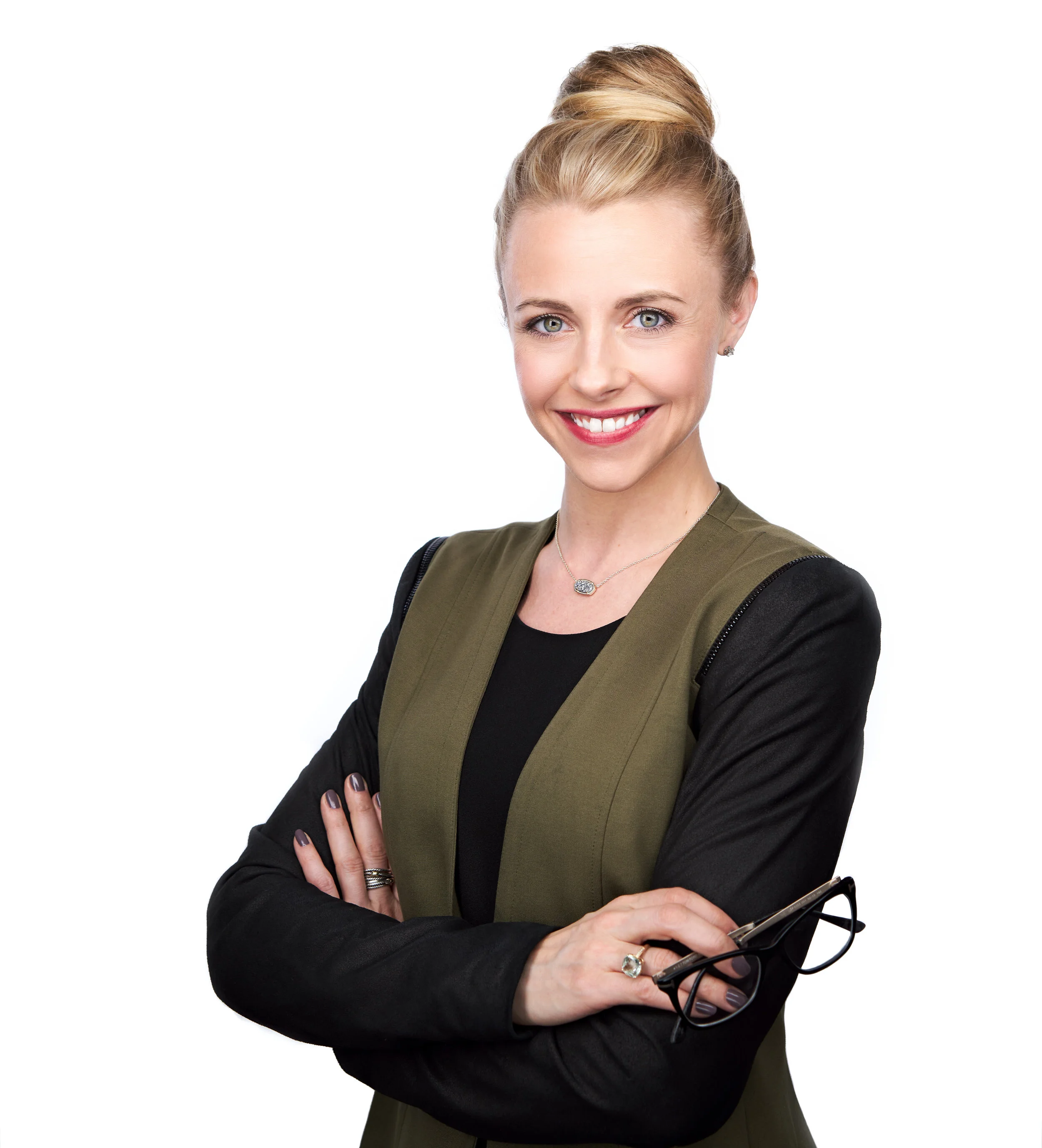 A confident woman with blonde hair styled in a bun, wearing a black top with an olive green blazer, smiling with arms crossed, holding glasses, standing against a white background.