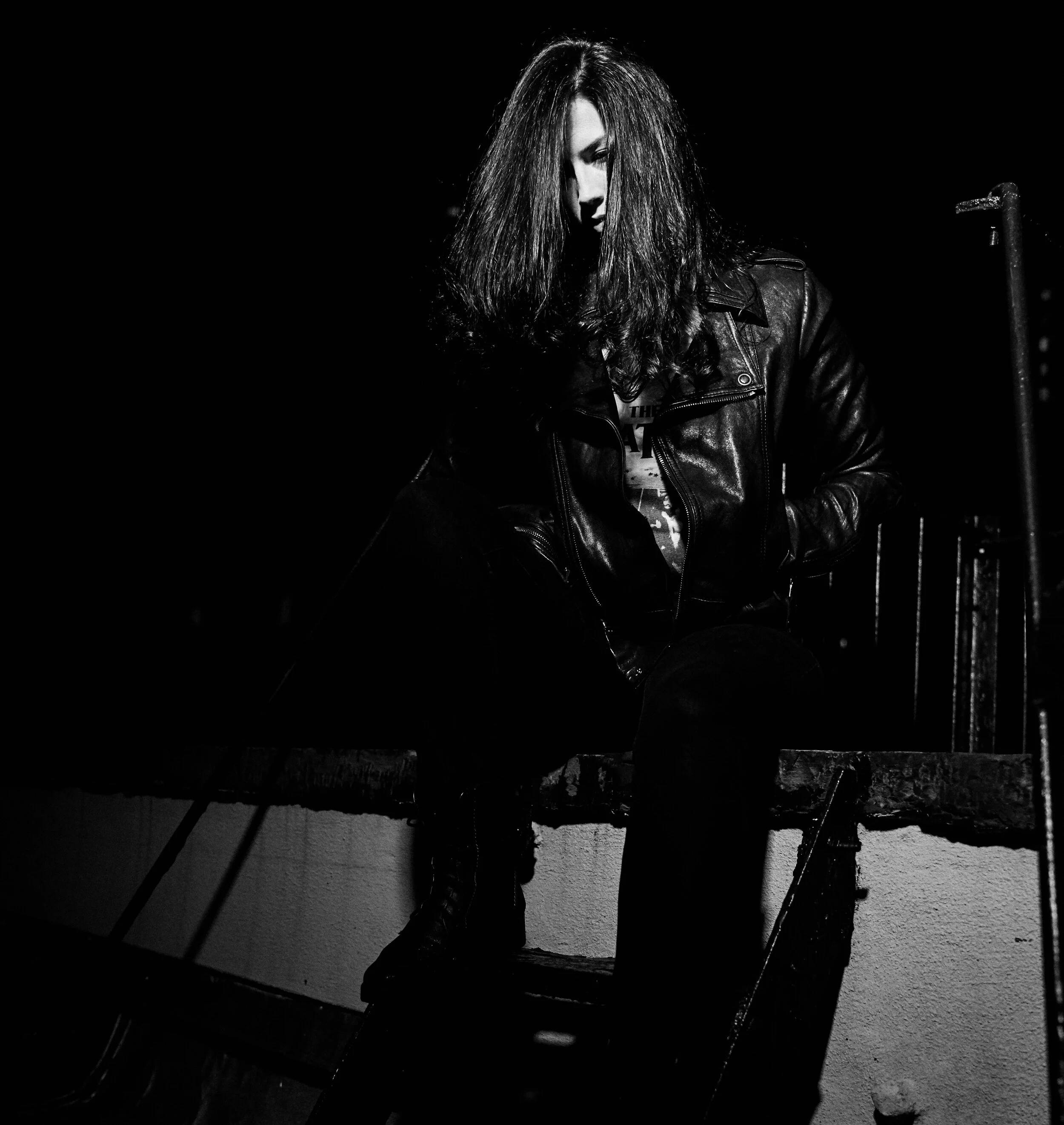 A woman with long, messy hair sitting on a wooden platform at night, wearing a leather jacket and dark clothing, with a dark background and low lighting.