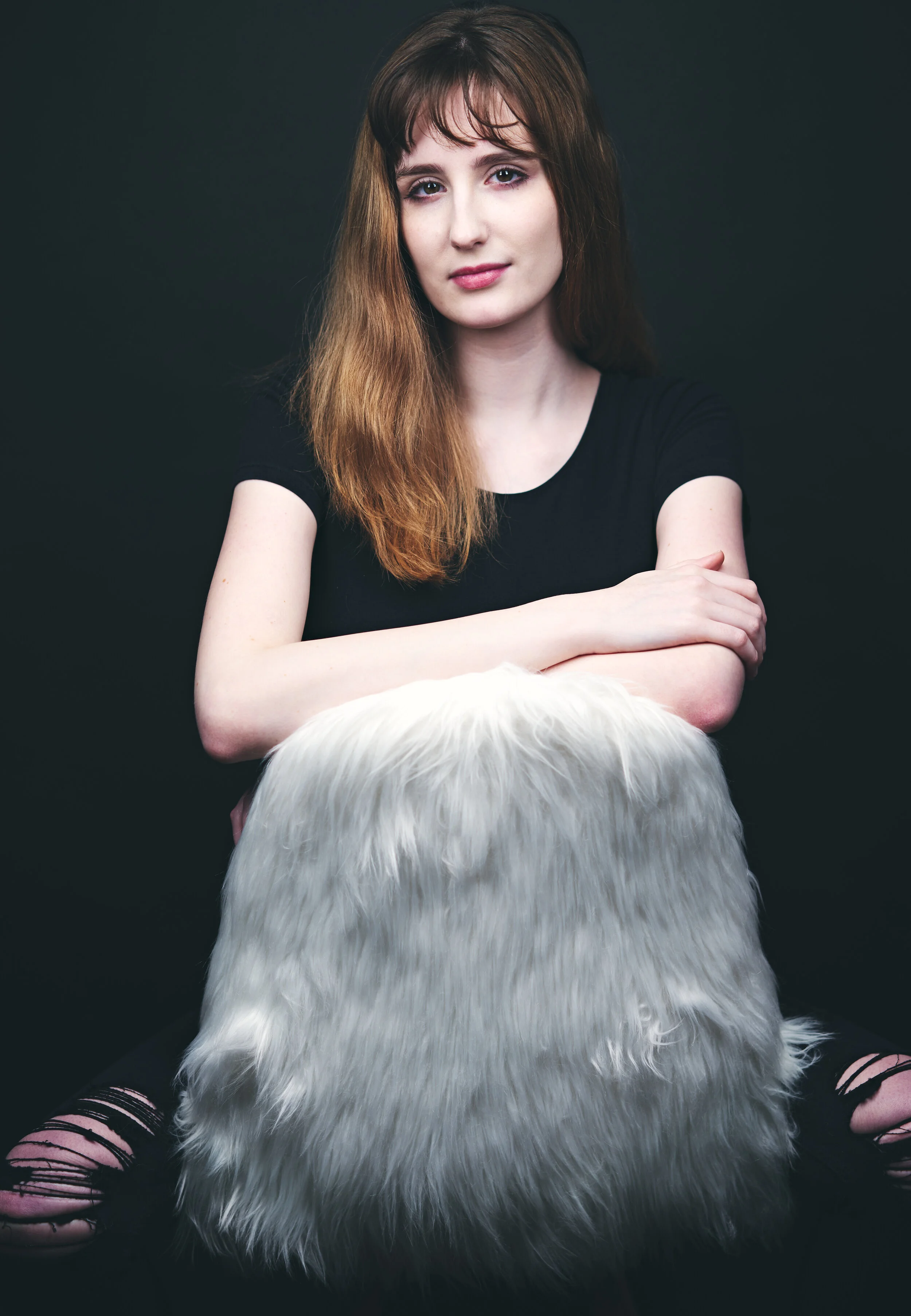 A woman with long brown hair wearing a black shirt sits with her arms crossed over a large white furry pillow in front of a dark background.