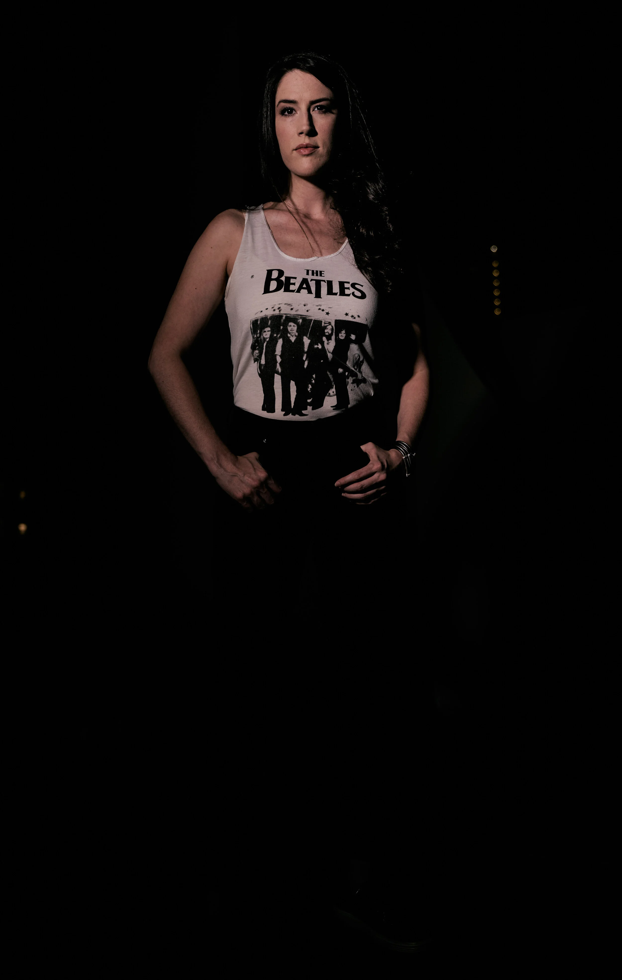 Woman wearing a white tank top with the Beatles logo and an image of the band, posing in a dark setting.