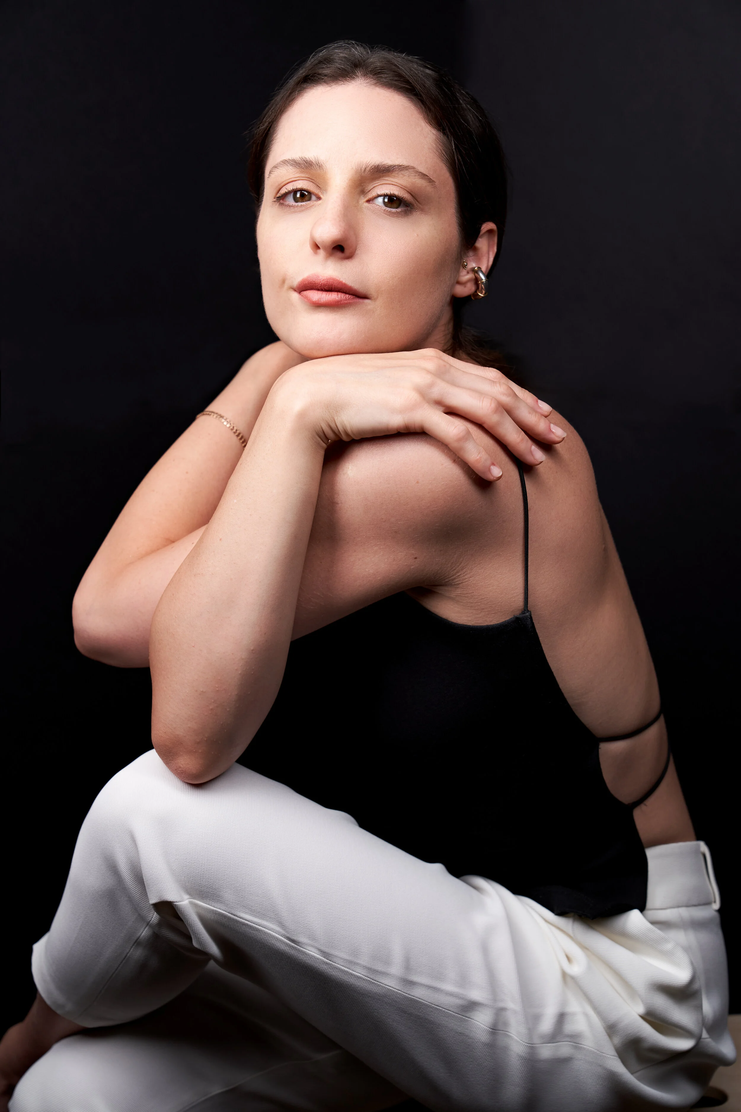 A young woman with dark hair in a braid, wearing a black spaghetti strap top and white pants, poses against a black background, resting her chin on her hand with a calm expression.