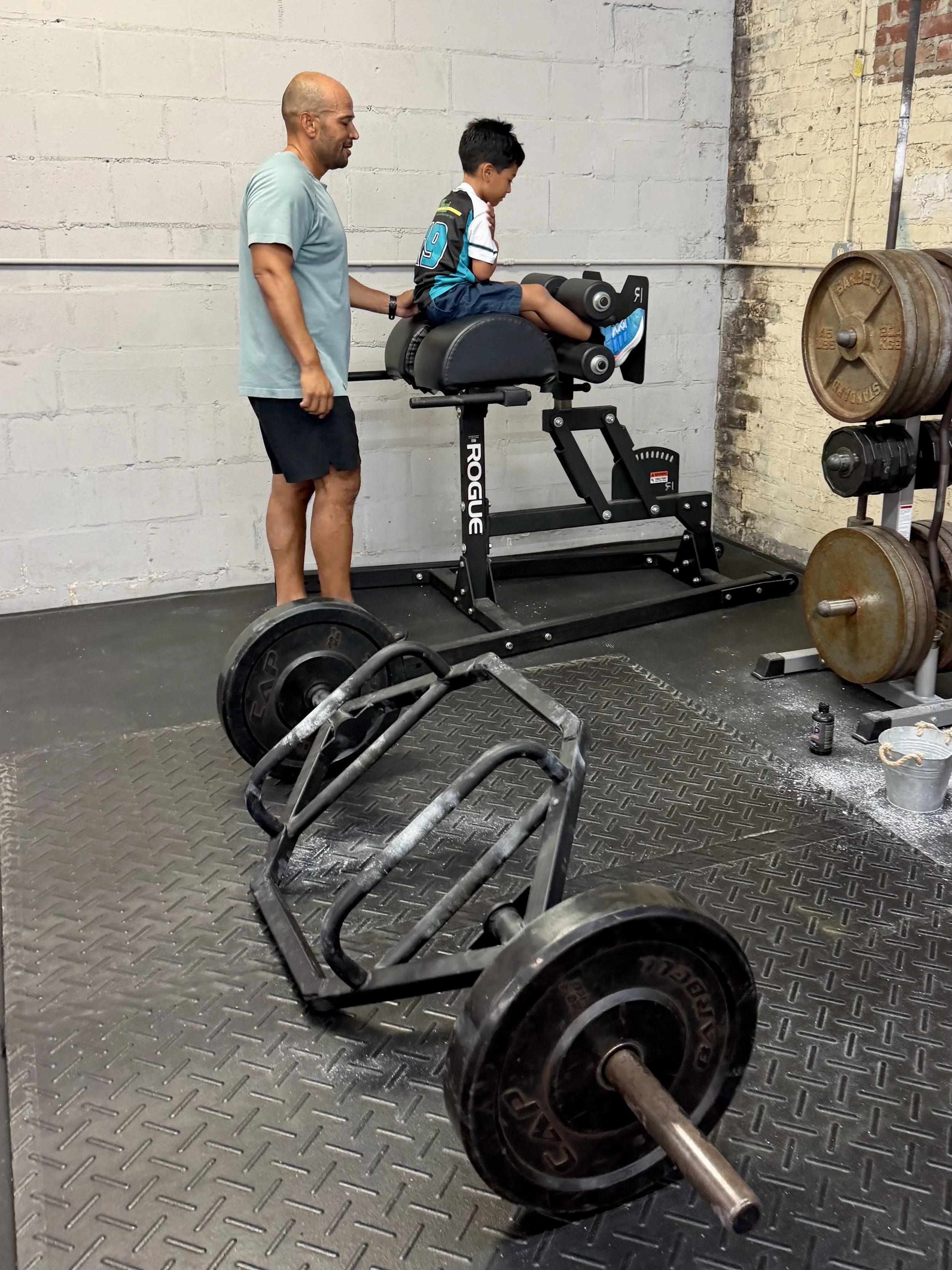 Perform24 member helping his 7-year-old son use a Rogue Glute Ham Developer during a summer workout as they build healthy family fitness habits together.
