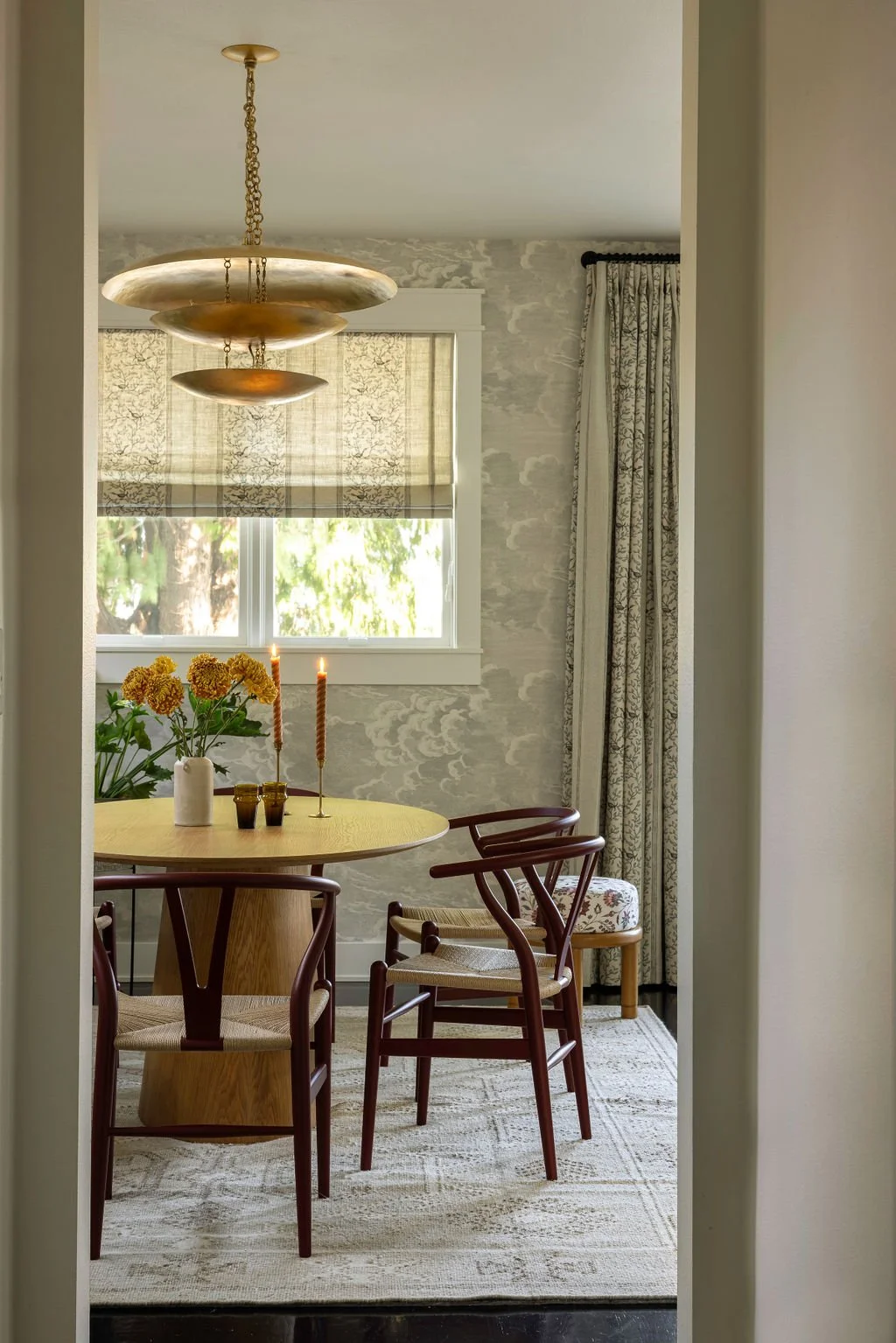 A cozy dining nook with a round wooden table, four chairs, a vase of yellow flowers, two lit candles, and a window with patterned curtains and a roller shade in a bright, decorated room.