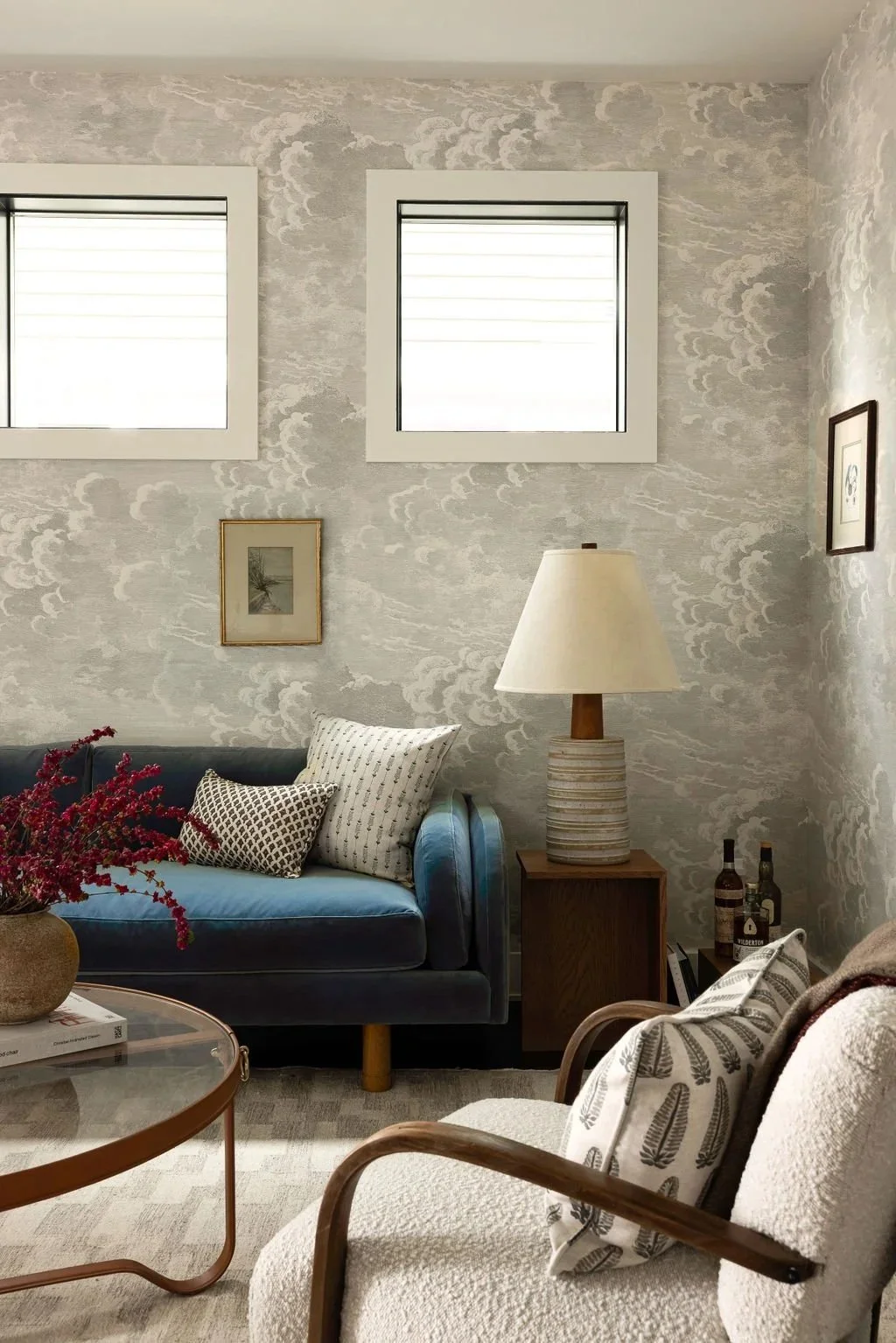 A cozy living room corner with a blue sofa adorned with patterned pillows, a wooden side table with a ceramic lamp, and a beige armchair with a patterned pillow. There are two framed pictures on the wall, and a small table with a vase of pink flowers, books, and bottles.