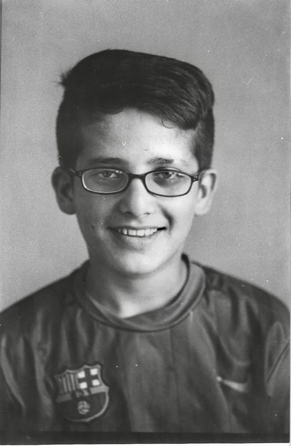 Black and white portrait photo of a young boy with glasses smiling, wearing a sports jersey with a crest on the chest.