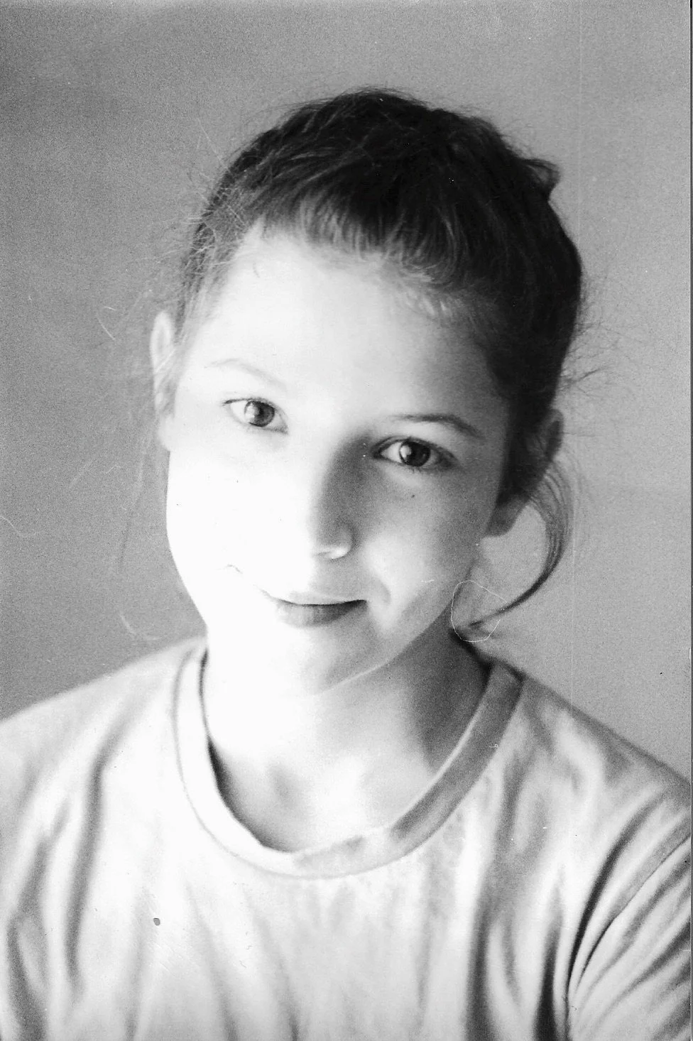 Black and white portrait of a young girl with light skin, smiling gently, wearing a t-shirt, with her hair pulled back and some loose strands framing her face.