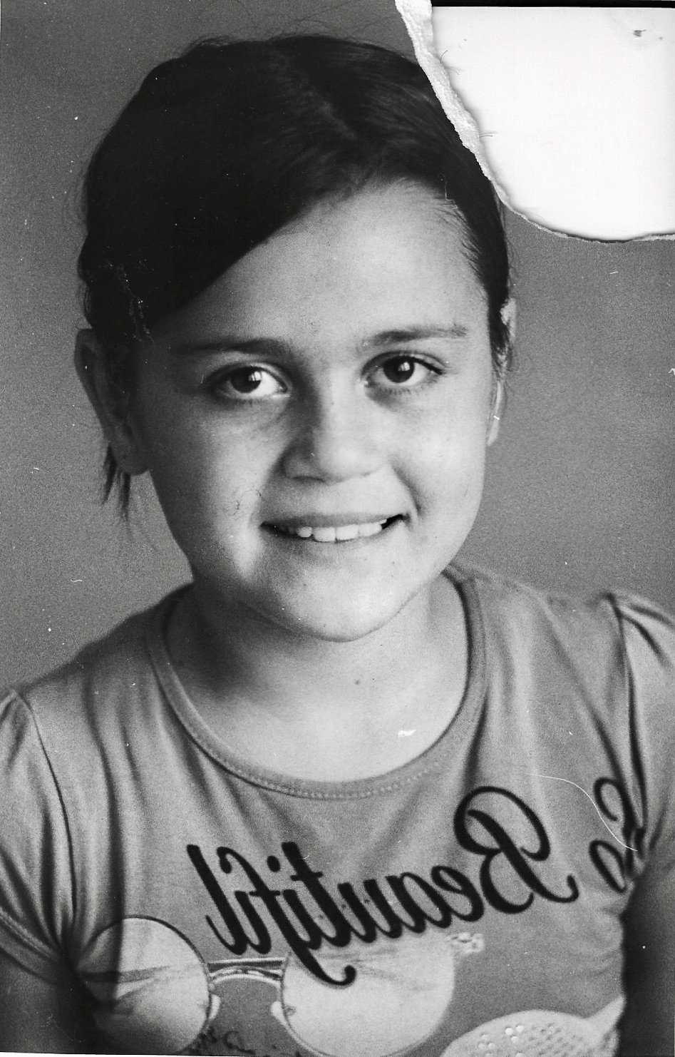 Black and white portrait of a young girl with short dark hair, smiling softly, wearing a T-shirt with text and graphics, with a torn piece of paper missing from the top right corner of the photo.