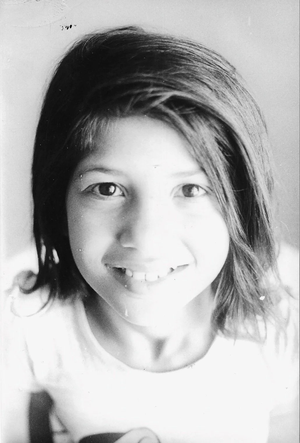 Black and white photograph of a young girl with shoulder-length hair, smiling and looking up at the camera. She is wearing a light-colored top.