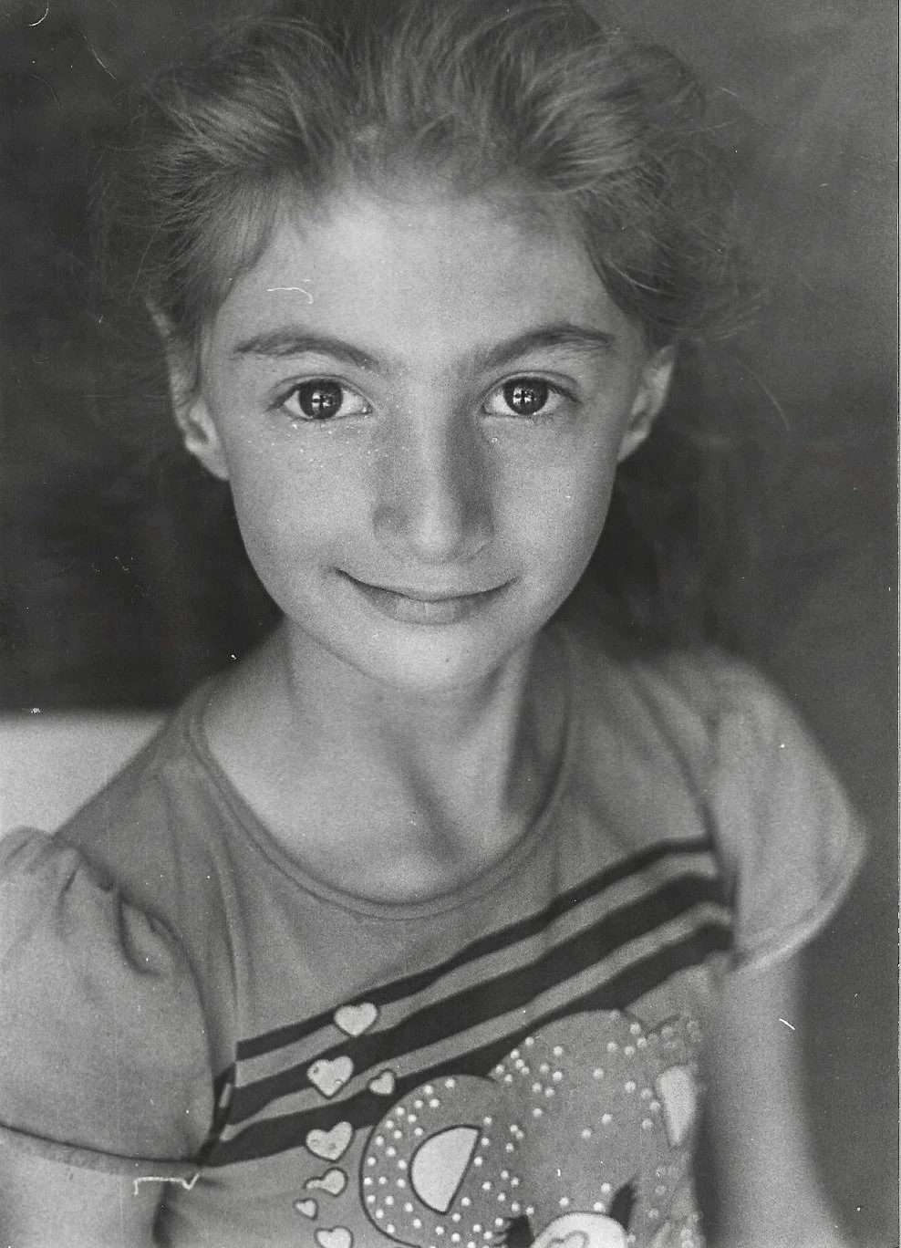 Black and white photograph of a young girl with curly hair, smiling, wearing a striped t-shirt with a decorative heart pattern.