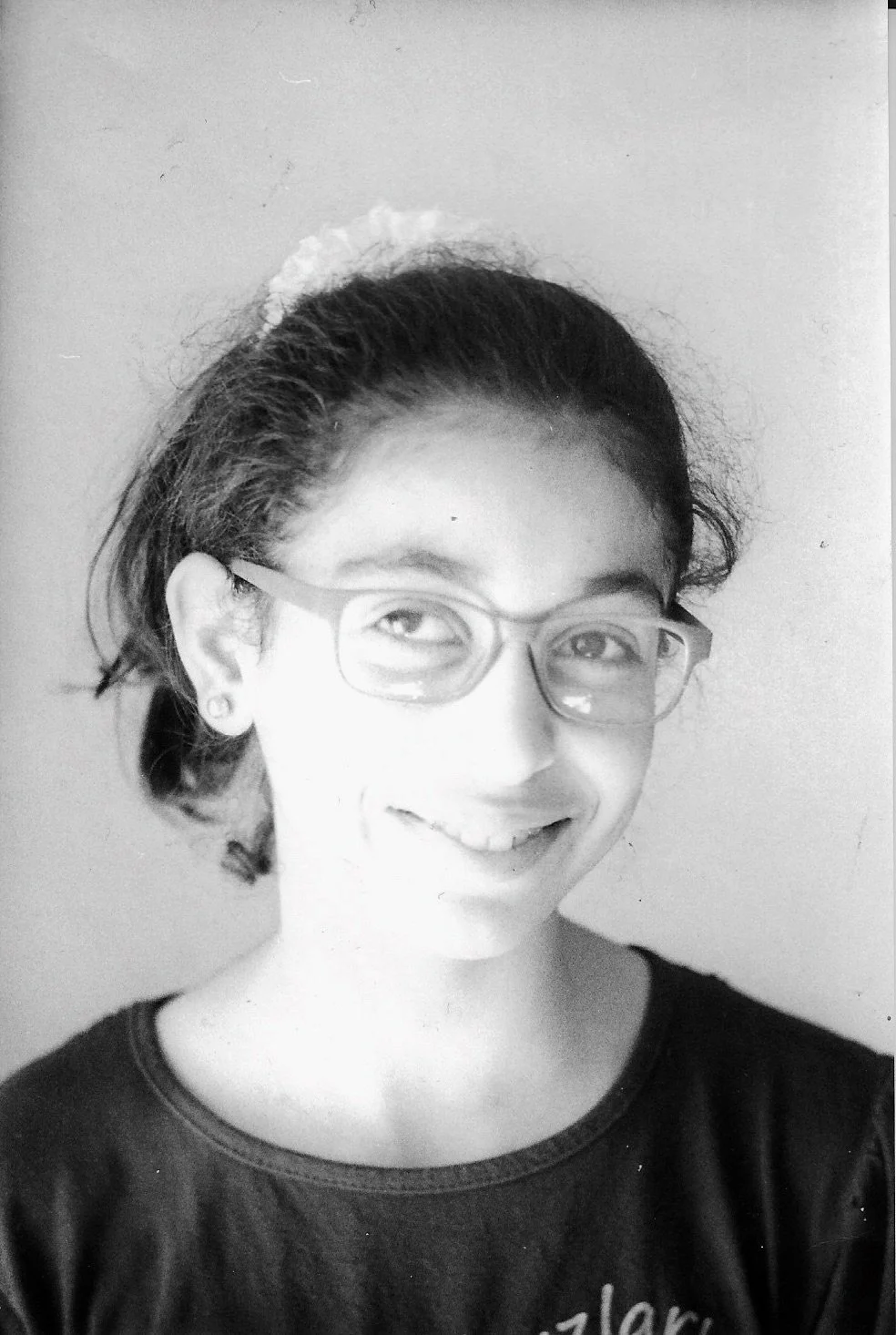 Black-and-white photo of a young girl with glasses, smiling, wearing a dark shirt, with hair pulled back and a headband or scrunchie.