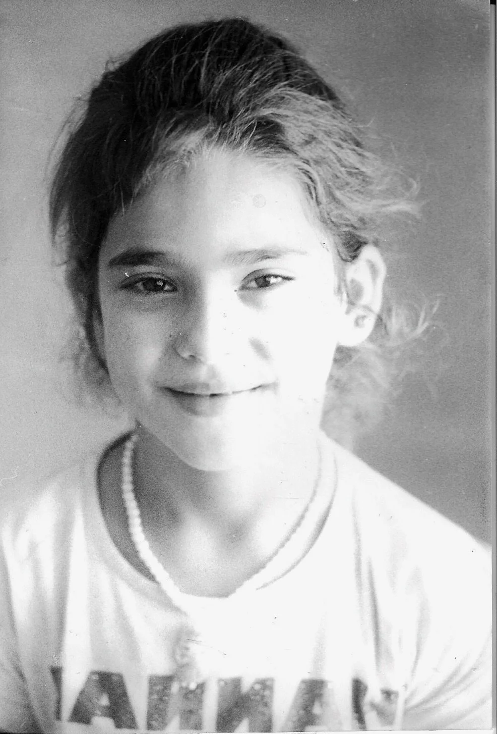Black and white portrait of a young person with short, tousled hair, wearing a pearl necklace and a shirt with visible text, smiling gently.