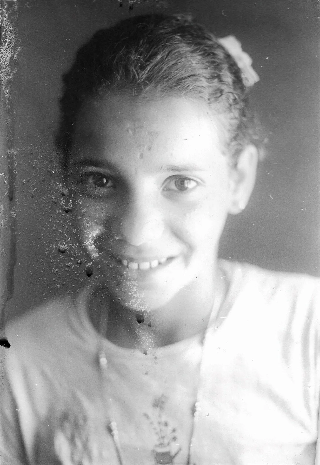 A black and white photo of a smiling young girl with curly hair, looking through a window with some dirt or scratches on it.
